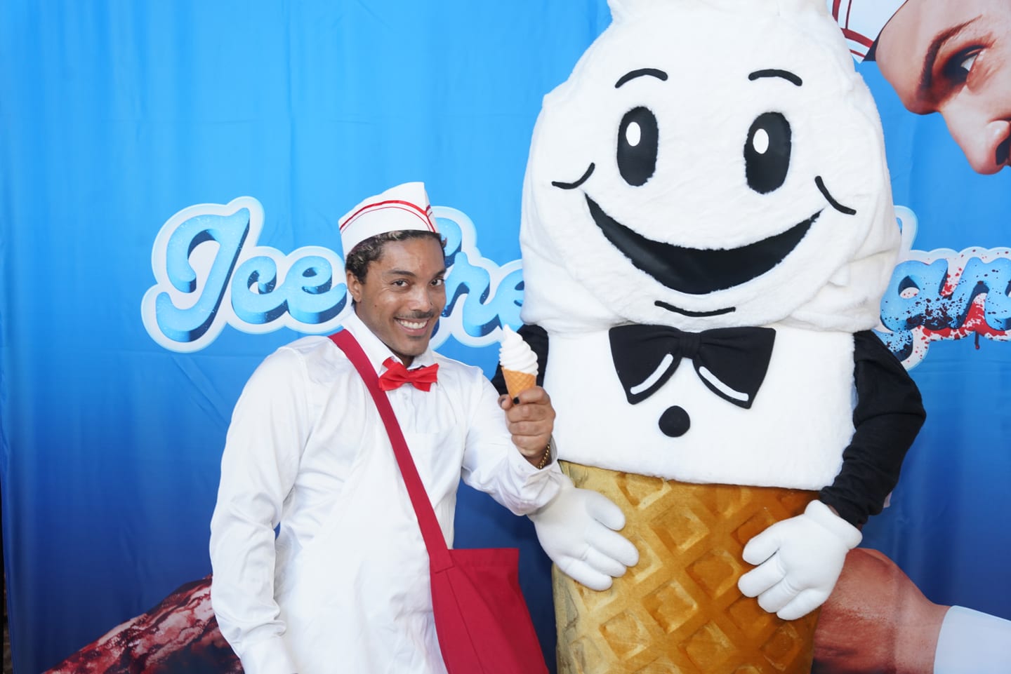 A costumed ice cream man character in a white uniform and red bow tie posing with a soft-serve cone mascot at the Ice Cream Man backdrop