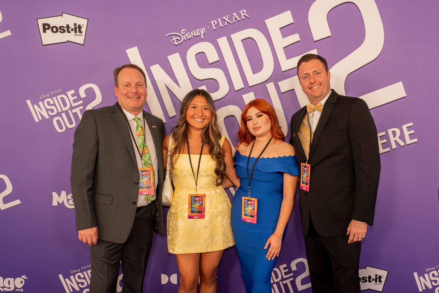 Four guests in formalwear with premiere credentials posed together on the purple Disney and Pixar Inside Out 2 World Premiere step-and-repeat, sponsor logos repeating across the wall behind them