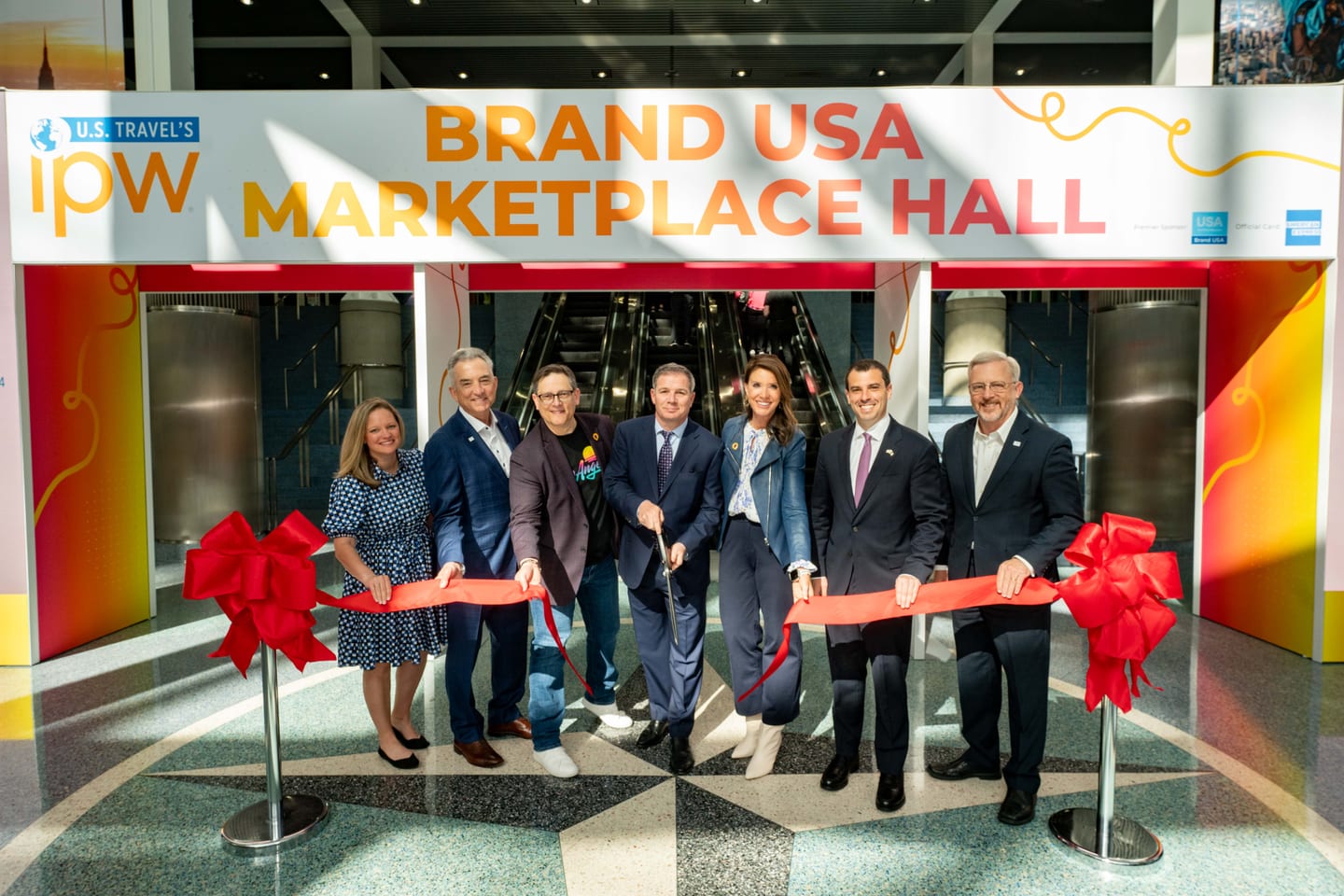 Seven travel-industry executives holding scissors to a red ribbon at the Brand USA Marketplace Hall entrance at IPW