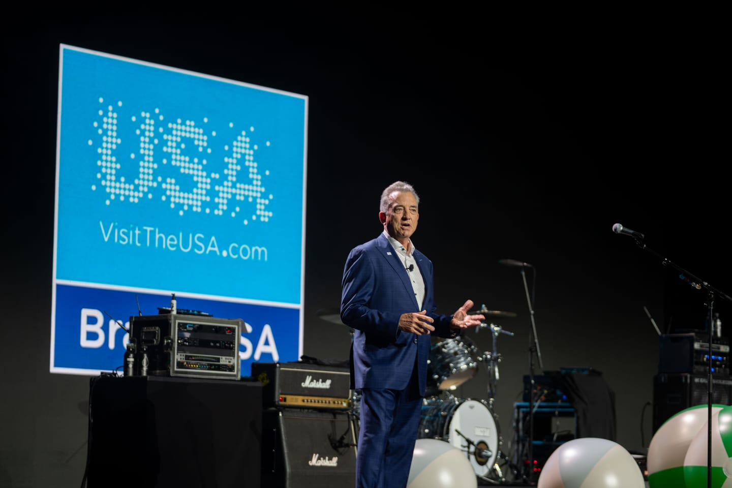 A speaker on stage beside a large Brand USA VisitTheUSA.com screen with band equipment set up behind him at IPW