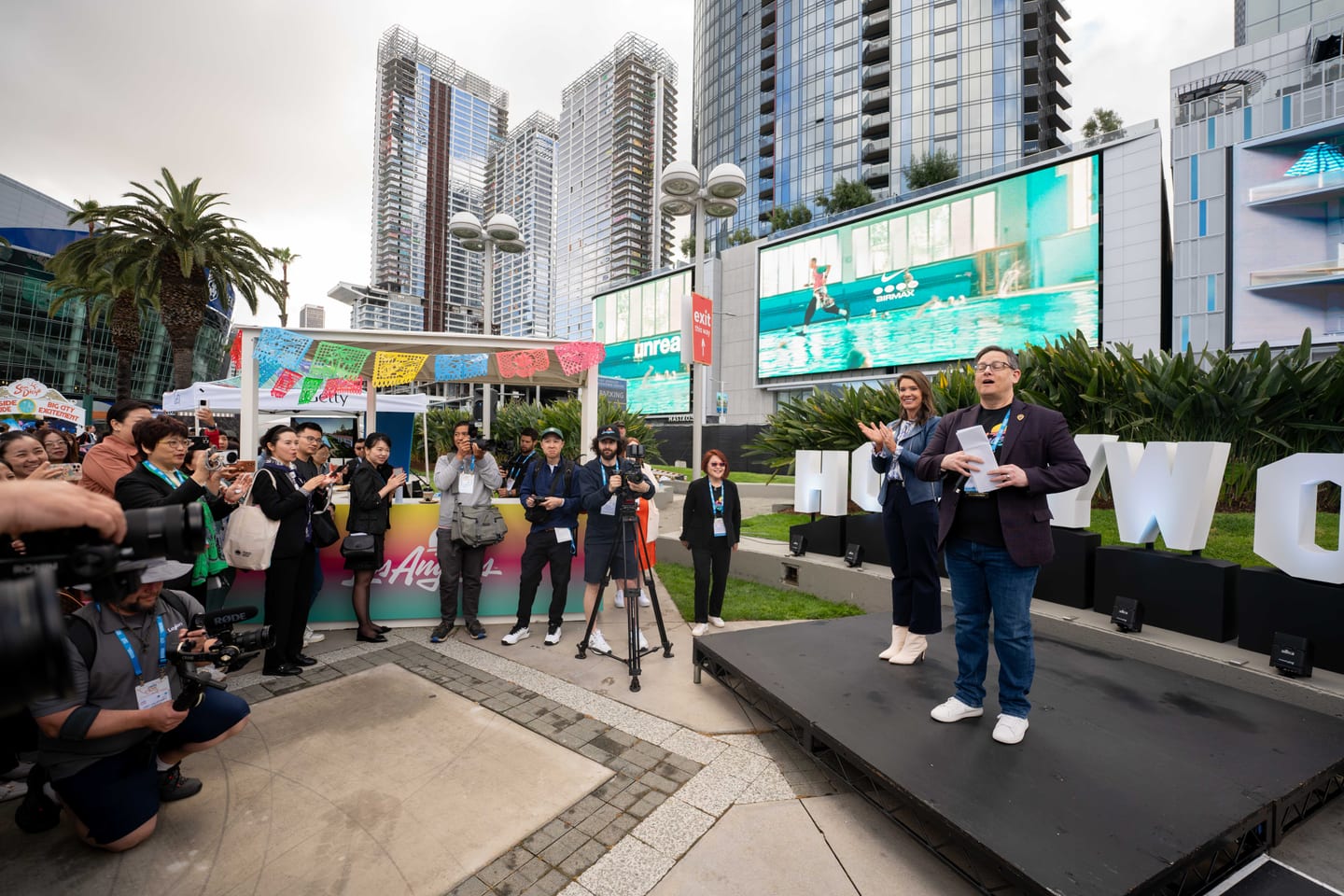 A host speaking on a small outdoor stage before press cameras and papel picado banners at IPW’s California Plaza