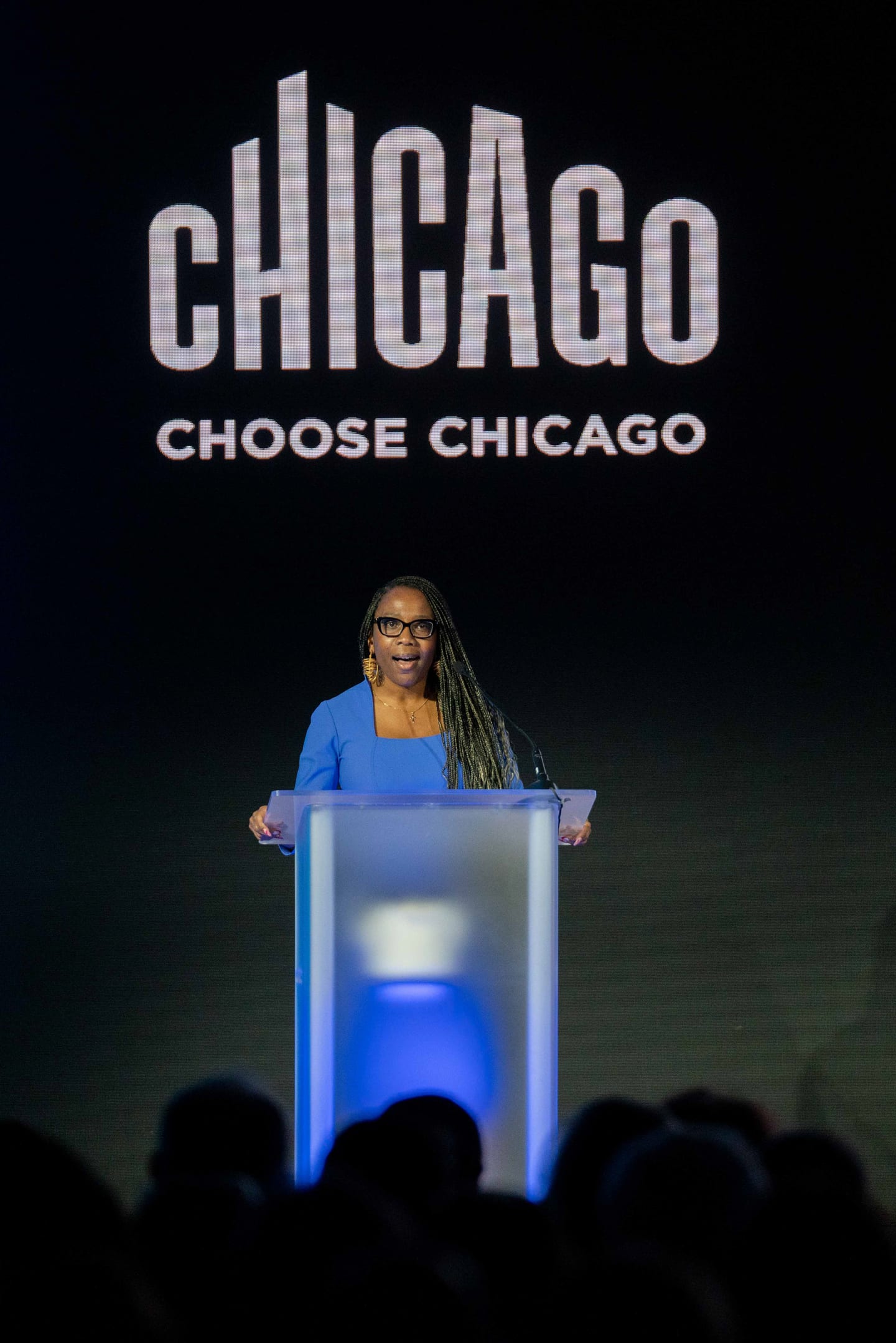 A speaker at a lit acrylic podium beneath a large Choose Chicago logo on a dark stage at IPW