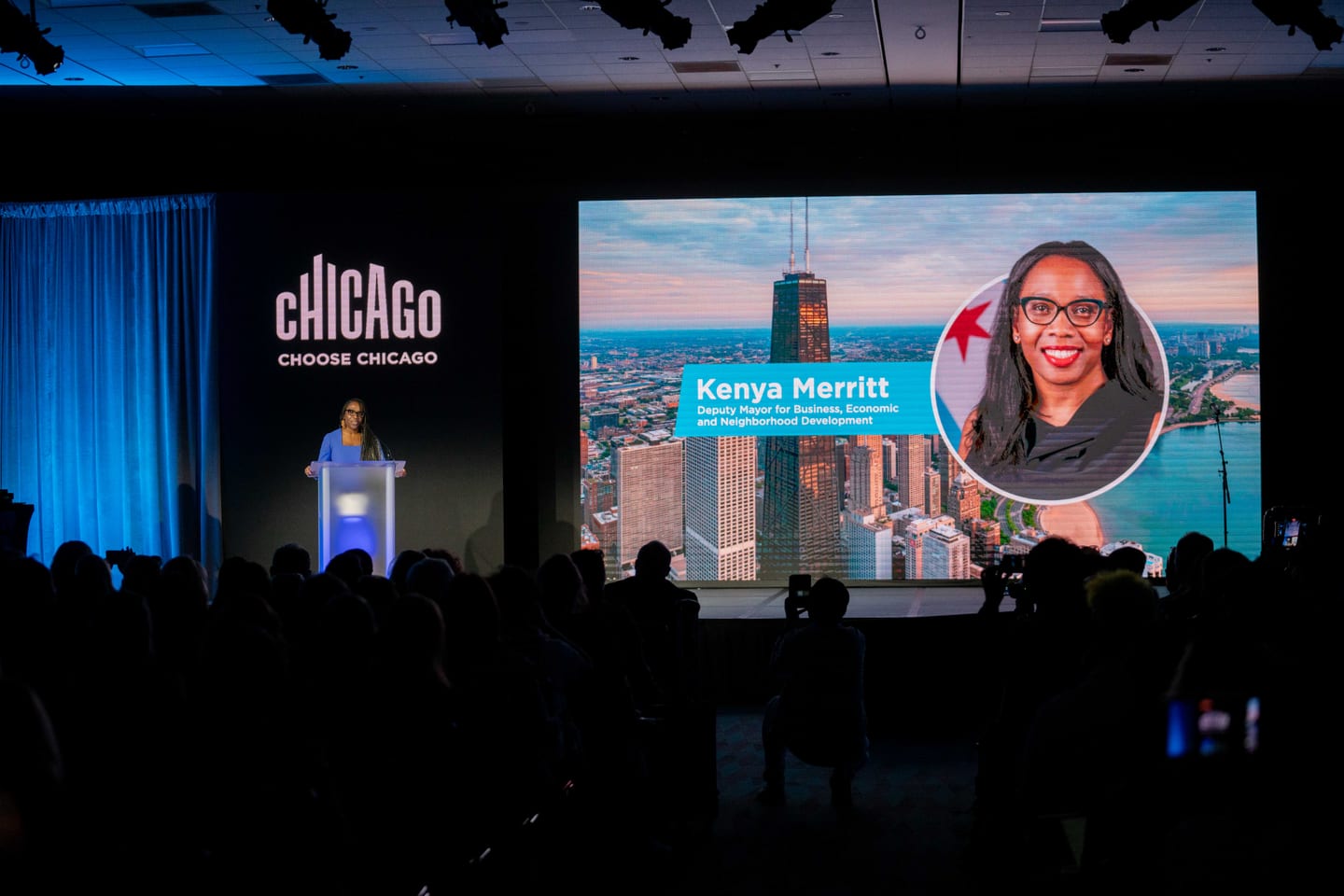 A speaker at a lit podium on the Choose Chicago stage as a Chicago deputy mayor is introduced on the screen behind at IPW