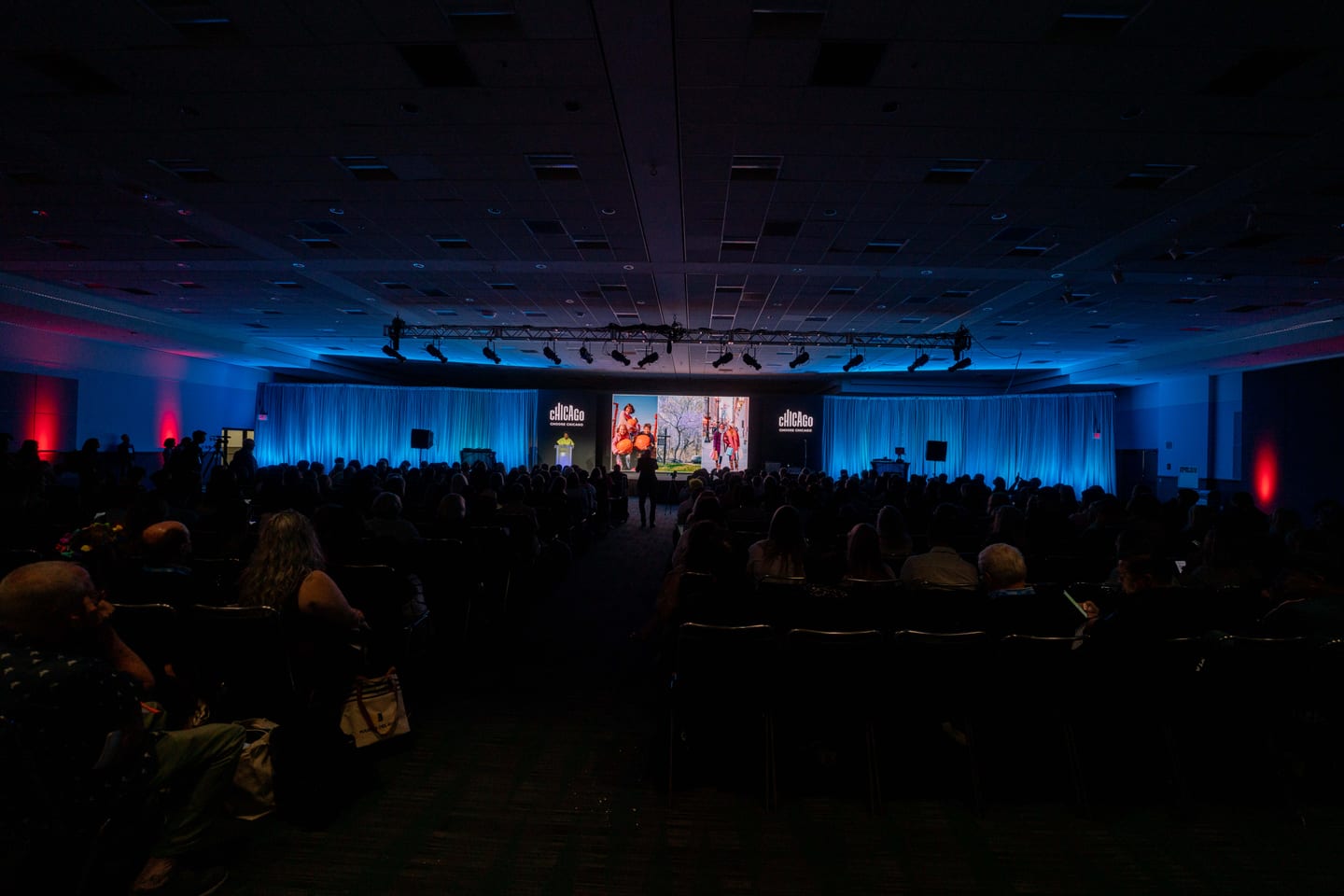 A wide view of a darkened IPW general session hall with a blue-lit stage and a seated audience