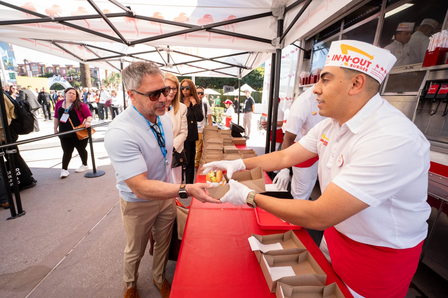 An In-N-Out server in a paper hat handing a burger across a red counter to an IPW delegate