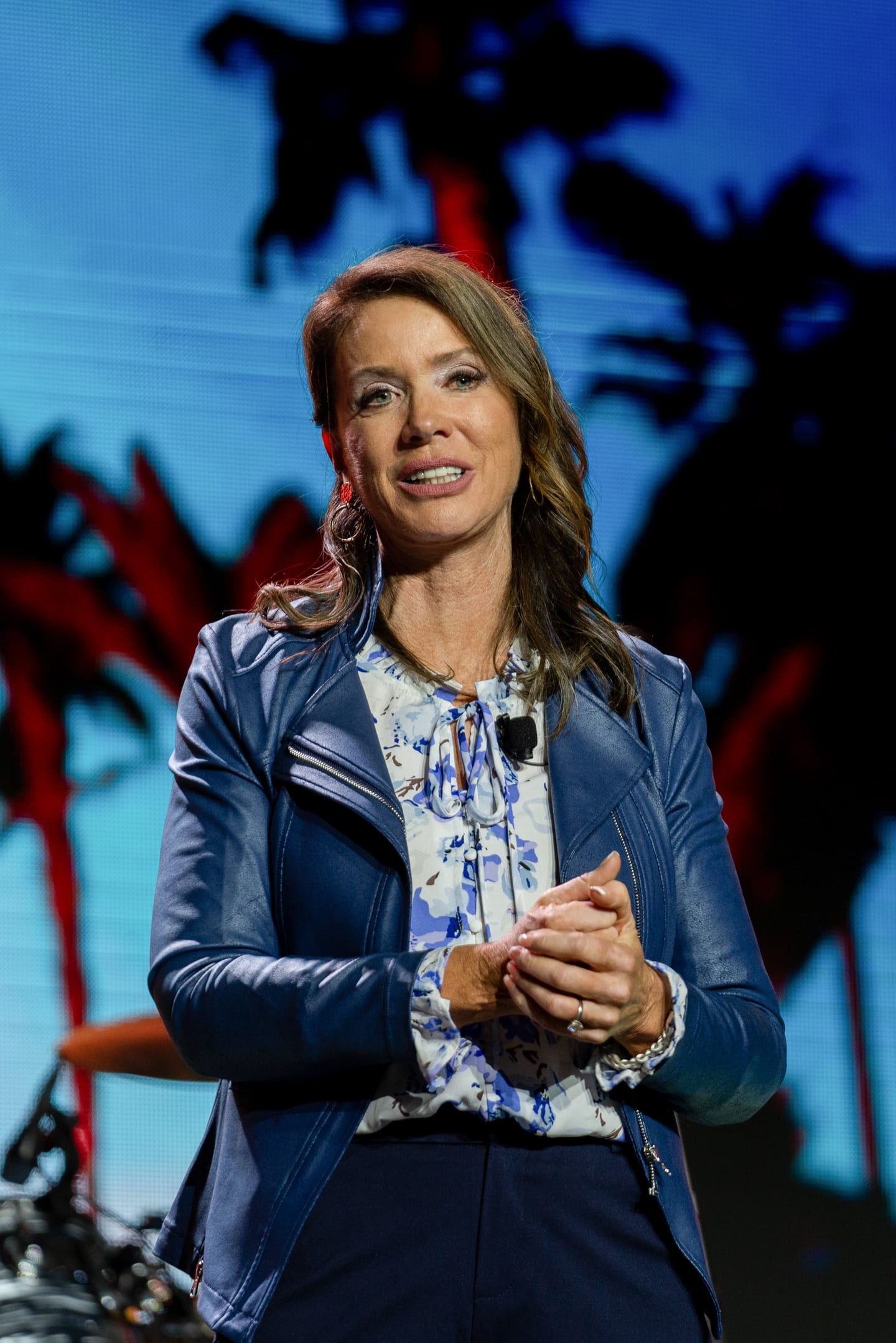 A speaker in a blue leather jacket mid-sentence on the IPW main stage against a palm-tree screen