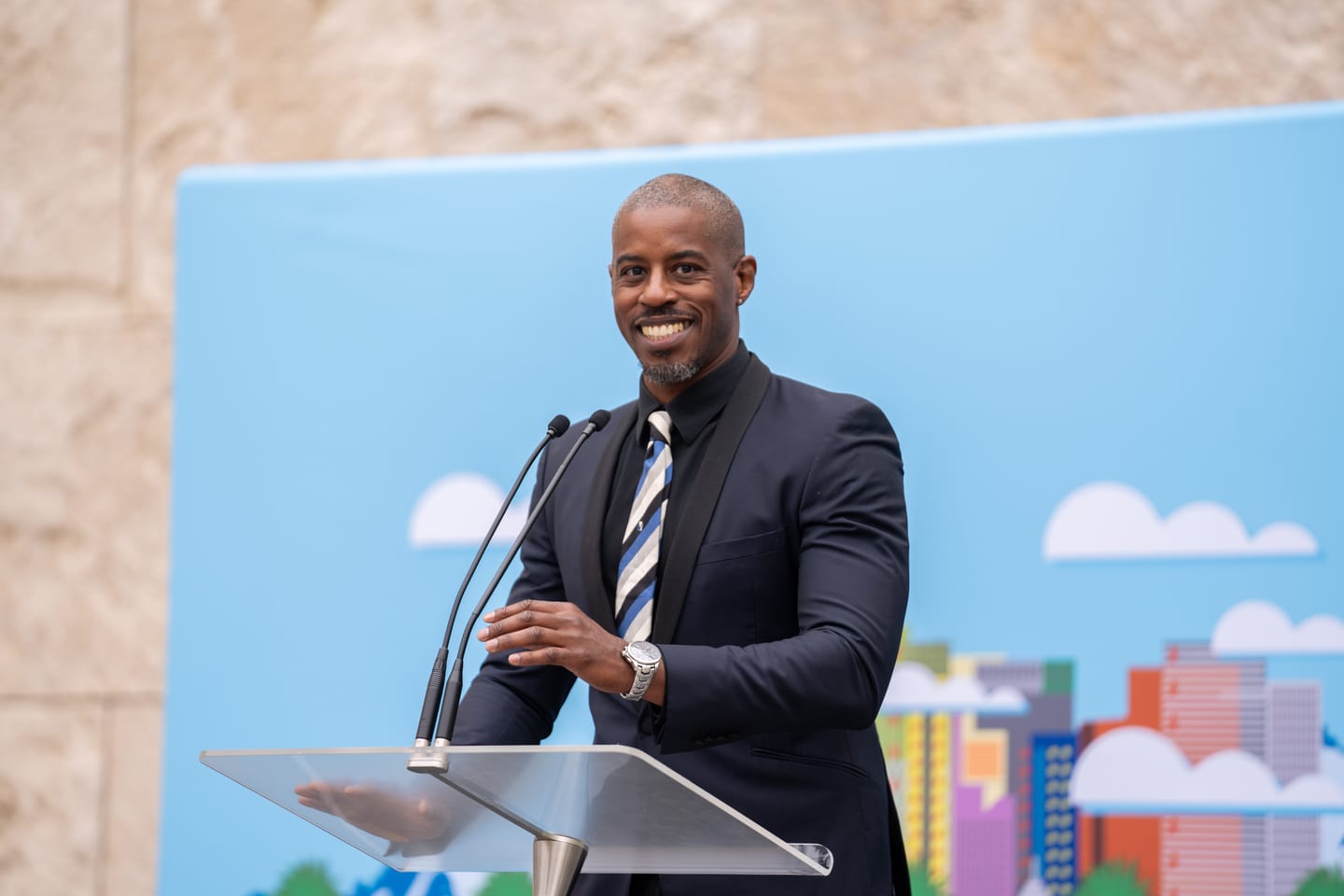 A speaker at a podium in front of a colorful Los Angeles skyline backdrop at an outdoor IPW welcome brunch