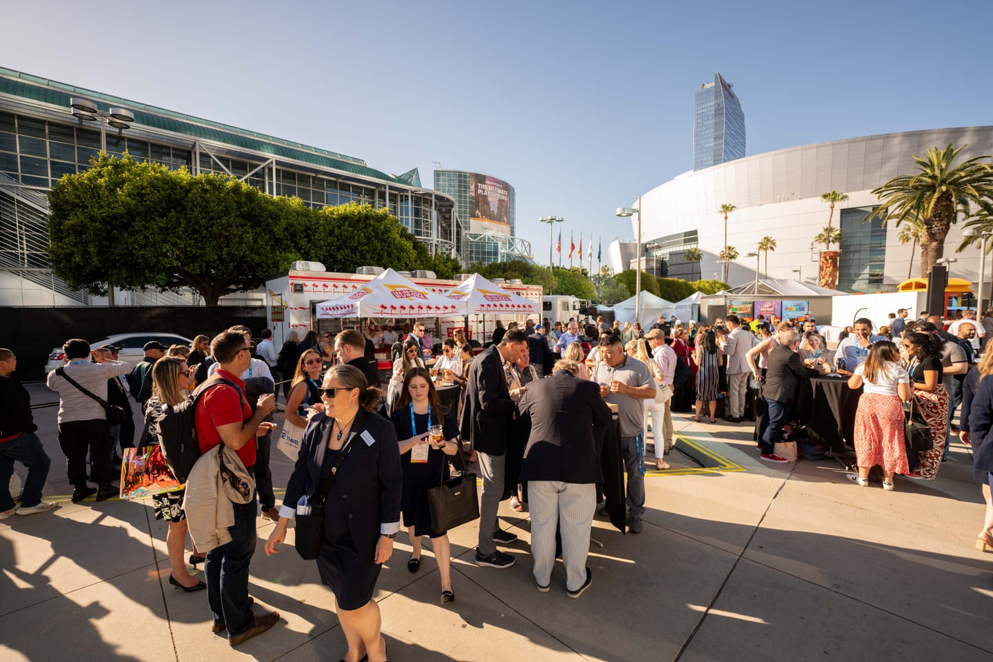 A crowd networking at an outdoor evening reception outside the Los Angeles Convention Center dome, with food trucks and string lights