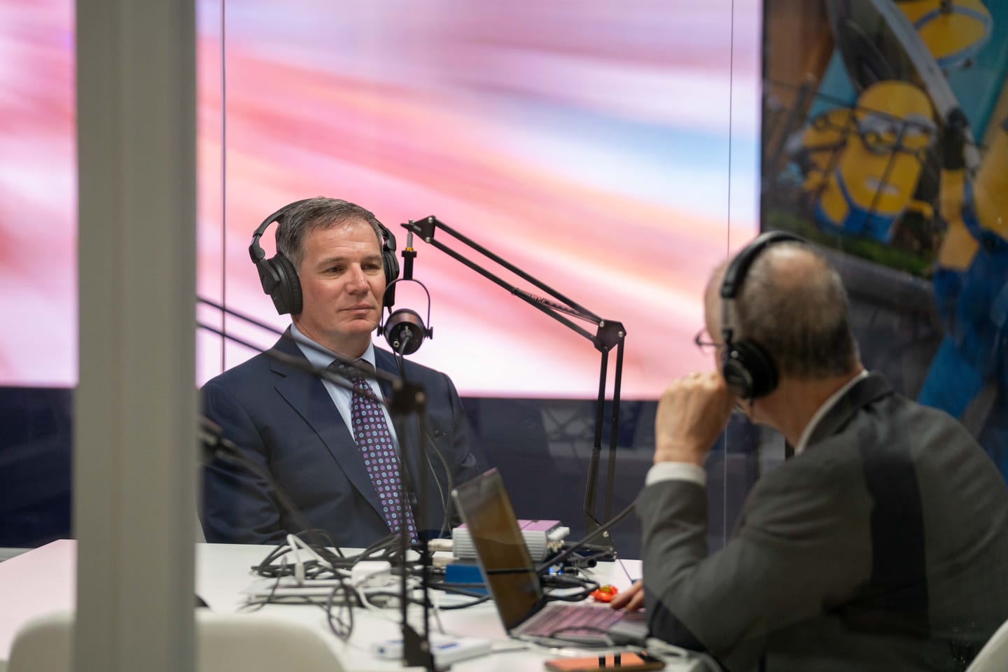 Two men in headphones recording a podcast interview at a broadcast desk in front of a bright screen at IPW