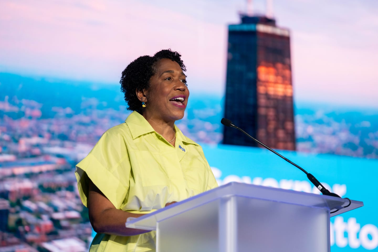 A speaker in a yellow jacket at a podium in front of a large city-skyline screen at IPW