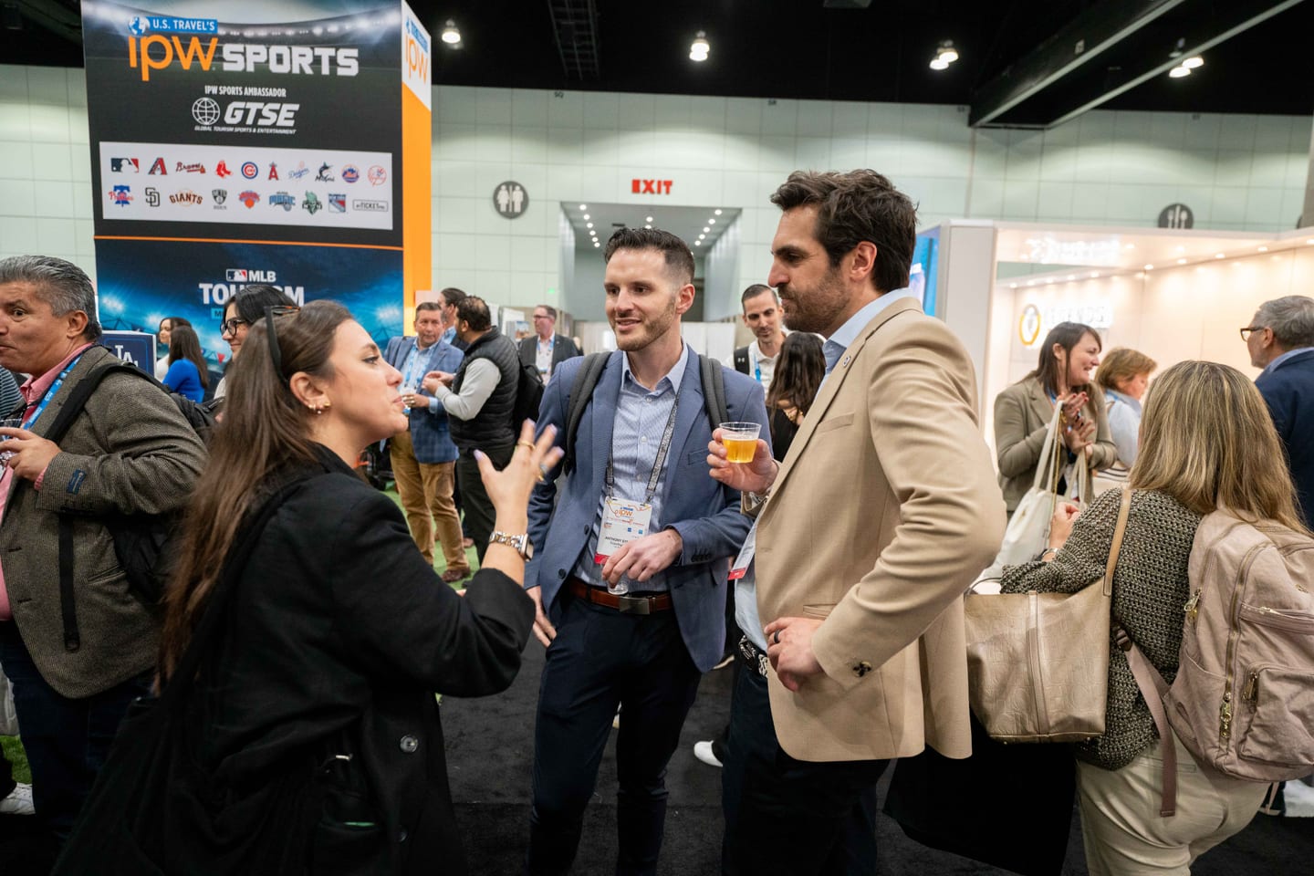 Delegates networking with drinks in front of an IPW Sports and GTSE booth displaying Major League Baseball team logos