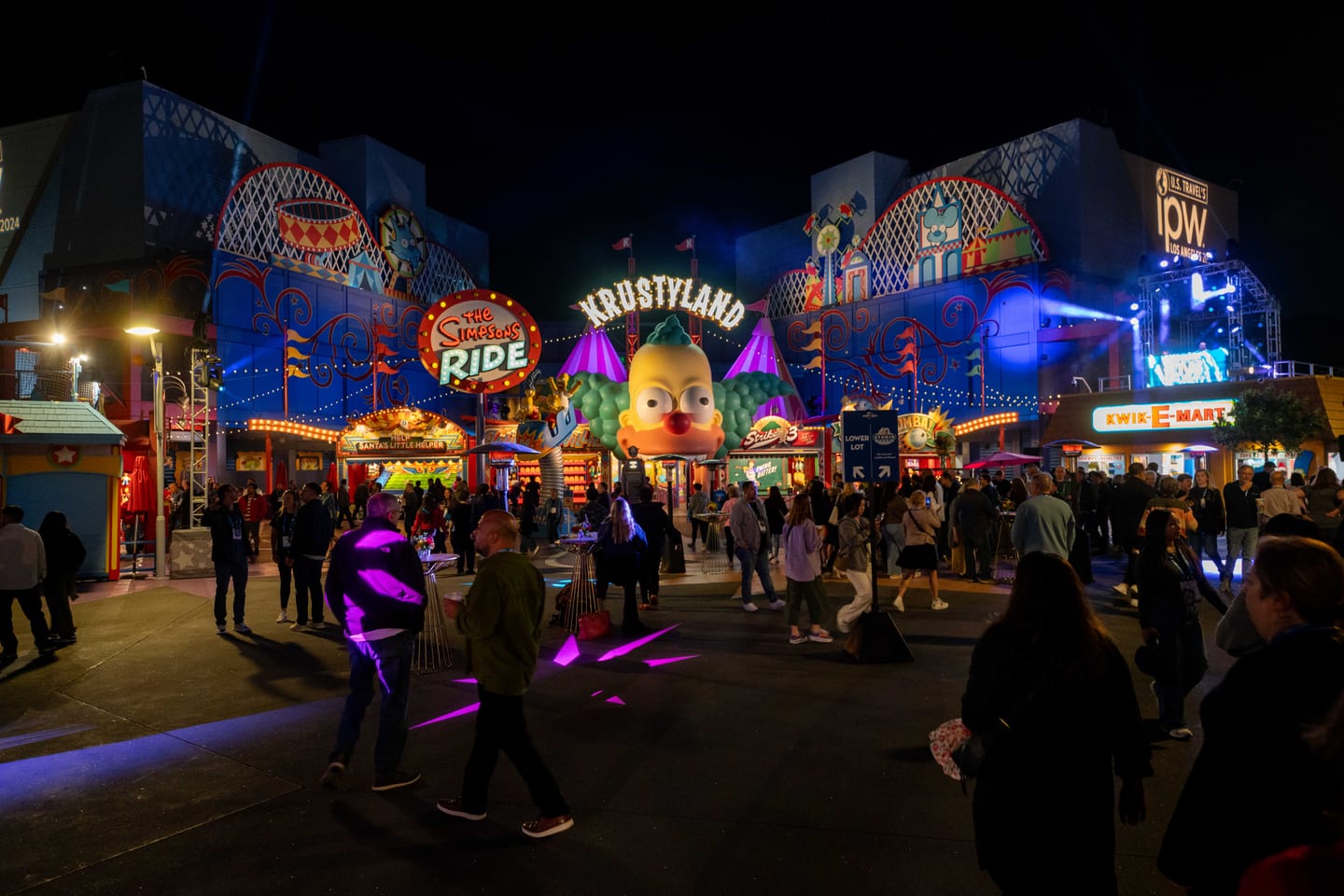 The Simpsons Krustyland area at Universal Studios Hollywood lit up at night with IPW branding and a crowd of delegates