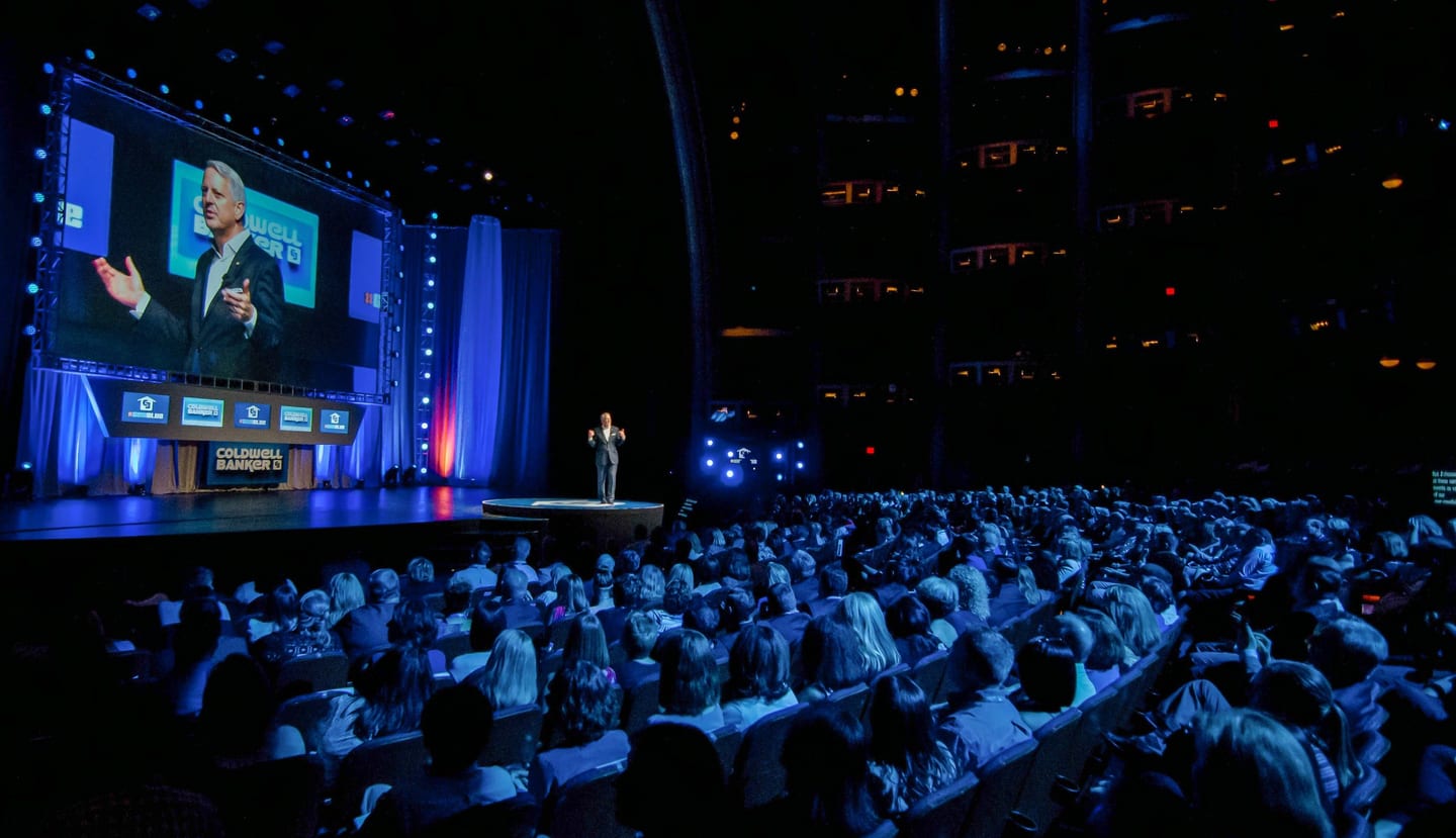 Wide view of a keynote stage and audience in a large darkened hall
