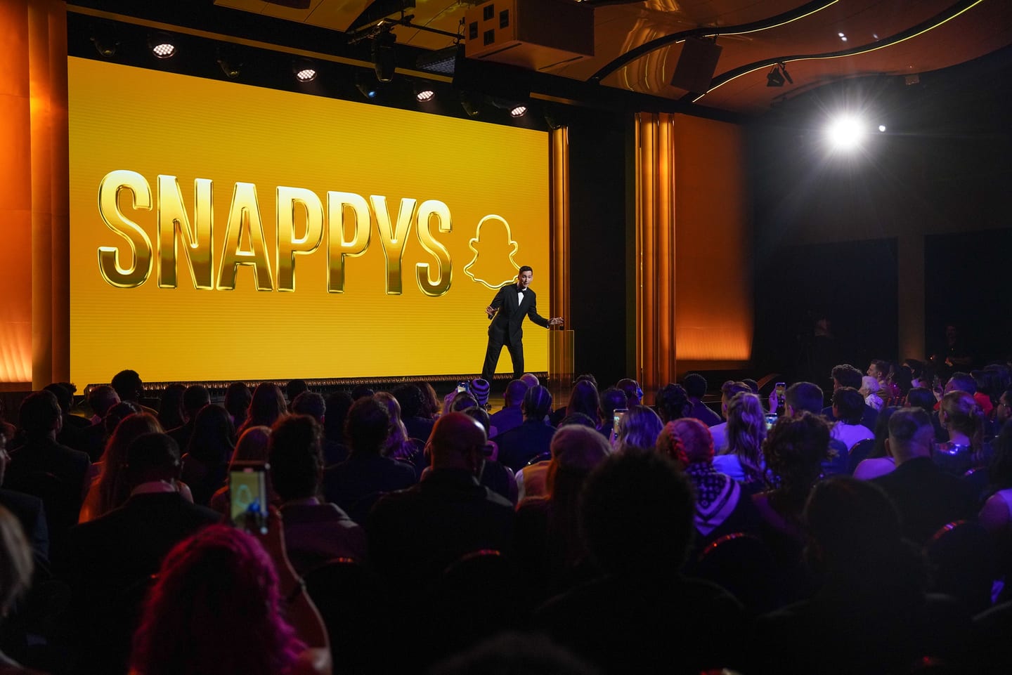 Comedian Matt Friend on stage in a tuxedo in front of a giant gold SNAPPYS screen at Snapchat’s creator awards in Santa Monica