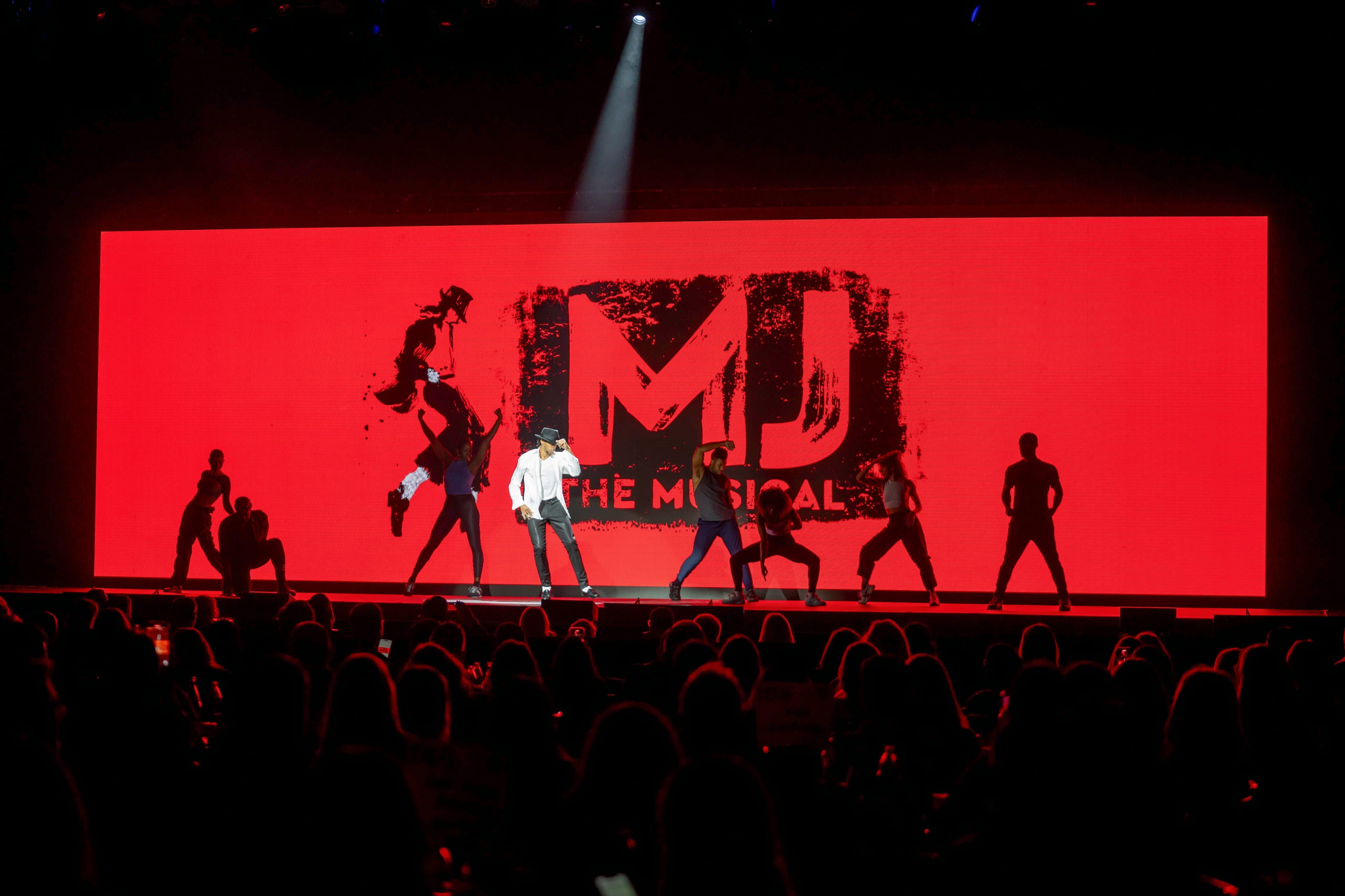 Dancers performing in silhouette on a stage against a red MJ The Musical LED backdrop under a spotlight, photographed over the heads of the audience