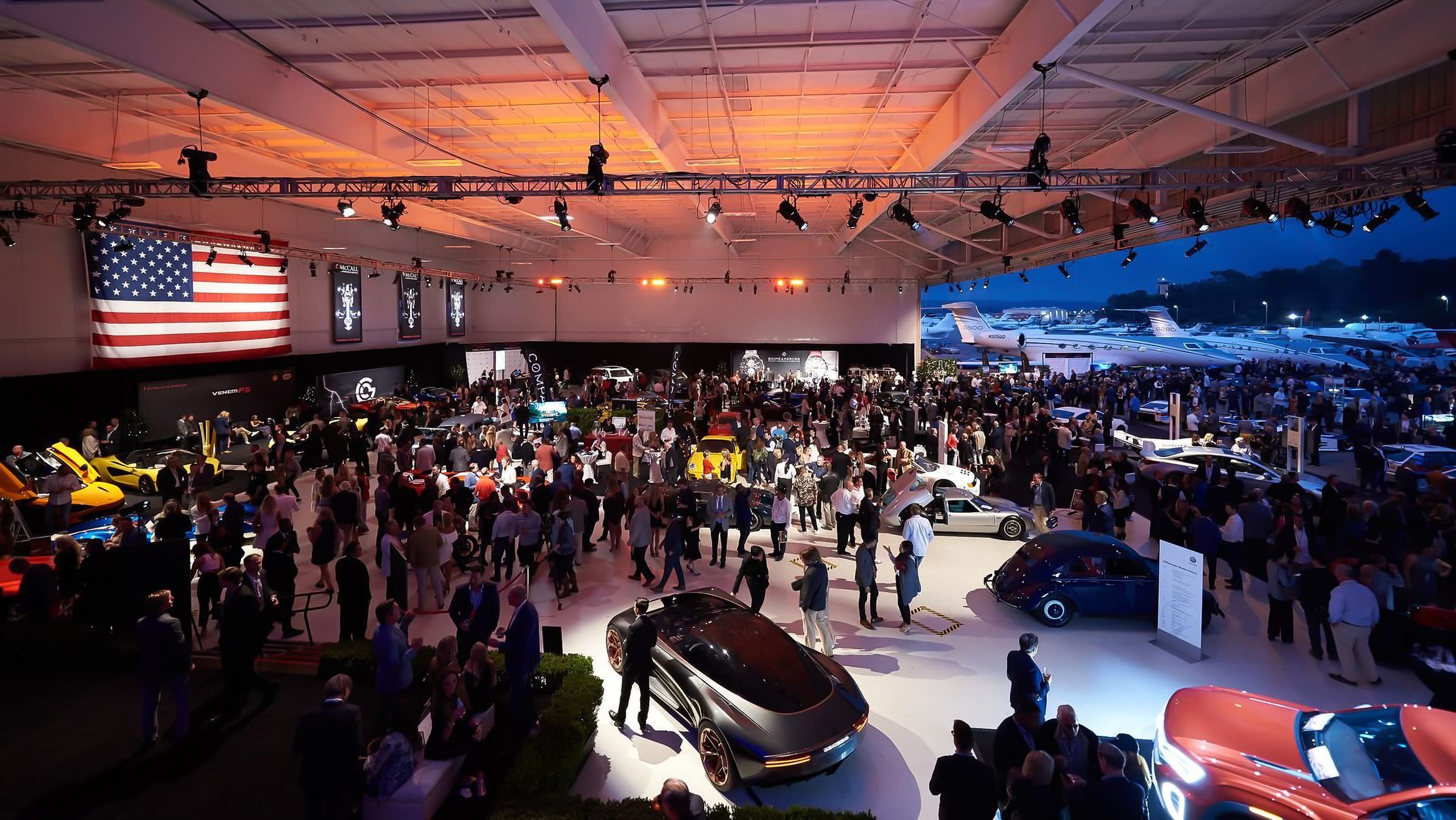 A packed automotive show floor inside a hangar at dusk with a large American flag on the back wall and aircraft visible through the open end at a Monterey Car Week event