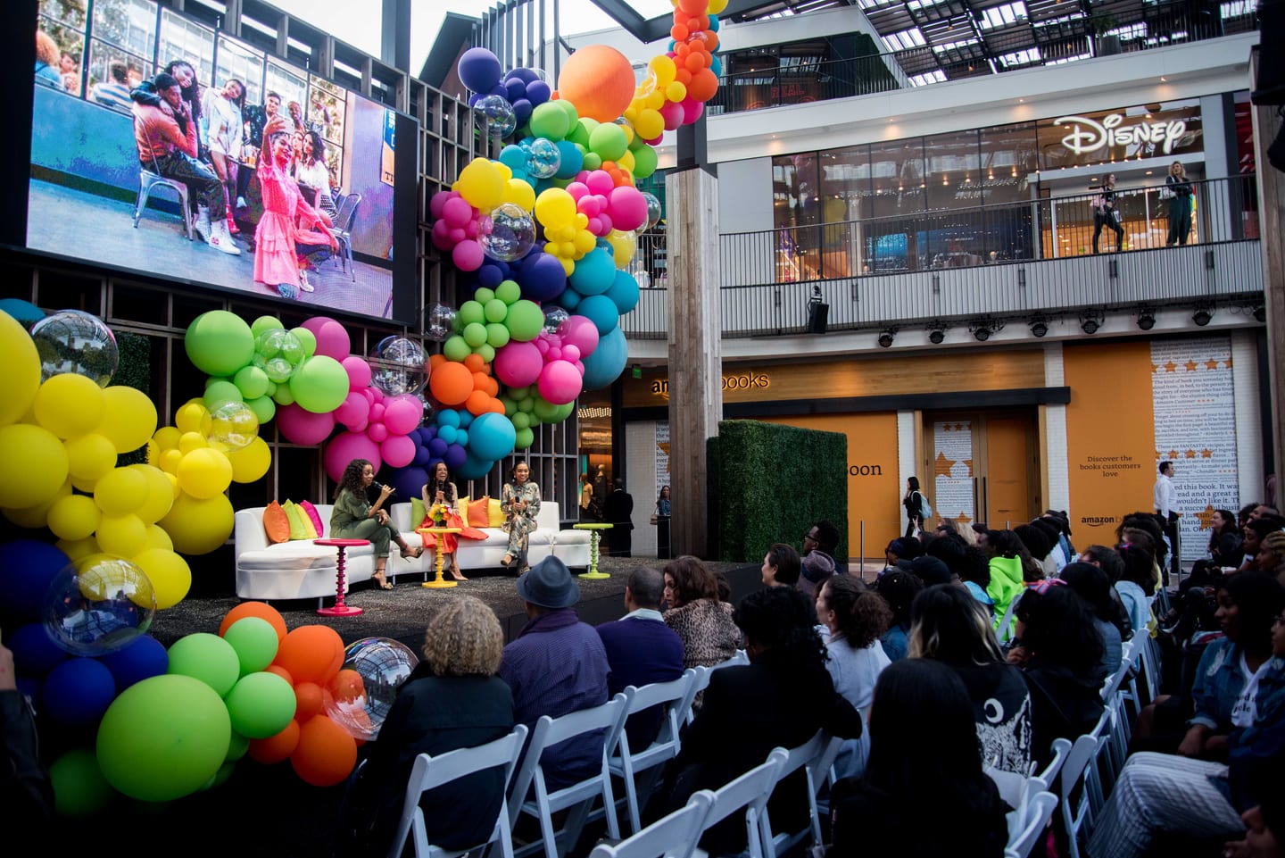 Three panelists with microphones seated on a white curved sofa on an outdoor stage framed by a huge rainbow balloon garland, a seated audience in front and a Disney store and Amazon Books on the balconies behind