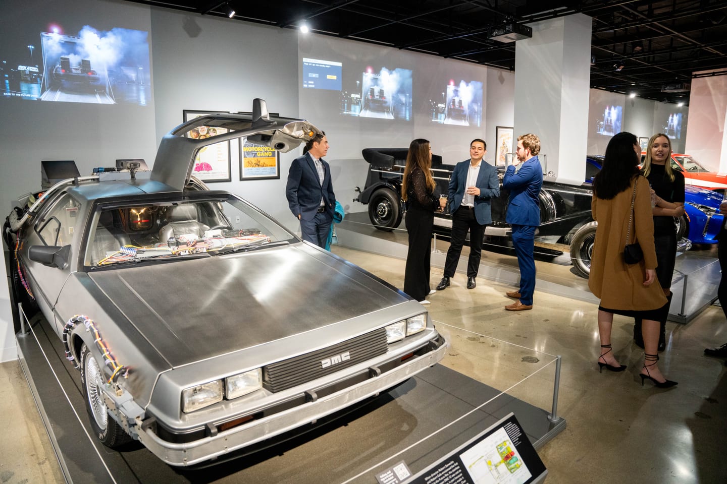 The Back to the Future DeLorean DMC-12 time machine with its gullwing door raised at the Petersen Automotive Museum, guests in suits networking in the gallery behind it
