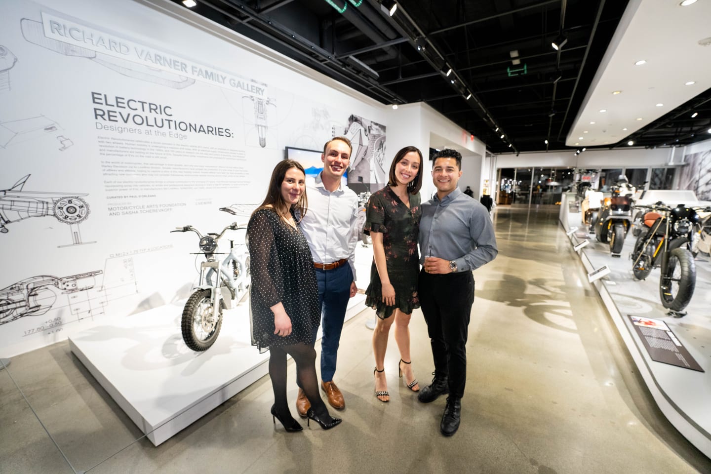 Four guests posed in front of the Electric Revolutionaries: Designers at the Edge exhibition wall in the Richard Varner Family Gallery at the Petersen Automotive Museum, electric motorcycles on display behind them