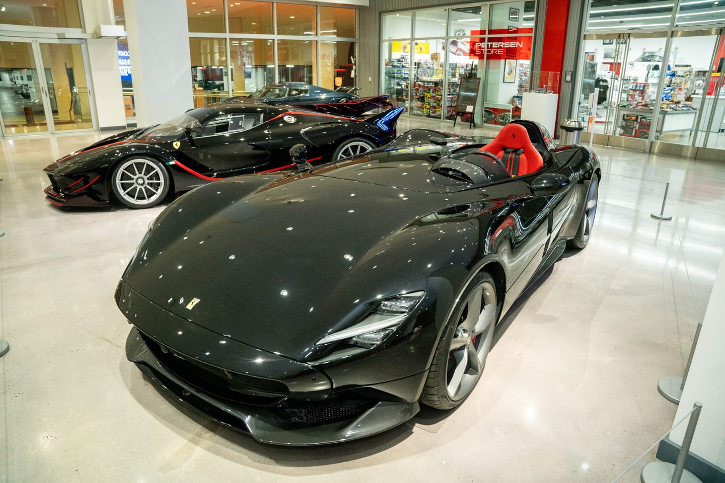 Two black Ferraris on the polished floor of the Petersen Automotive Museum lobby, an open-cockpit speedster with a red seat in the foreground and a striped hypercar behind it, the Petersen Store lit up in the background