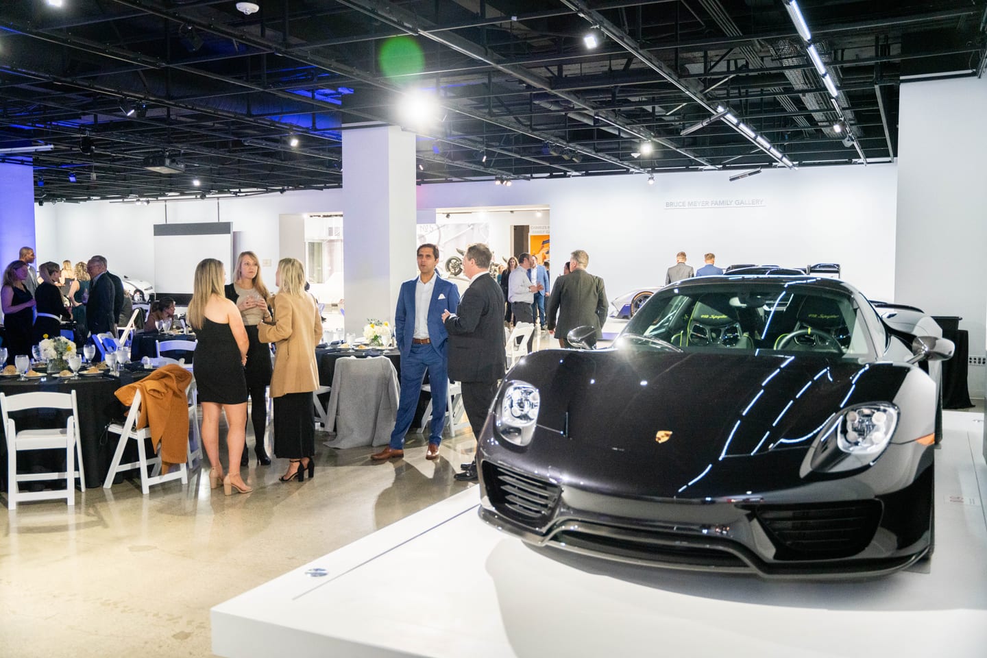 A black Porsche 918 Spyder on a white plinth in the Bruce Meyer Family Gallery at the Petersen Automotive Museum, guests in business attire talking beside set dinner tables at an FTI Consulting corporate reception