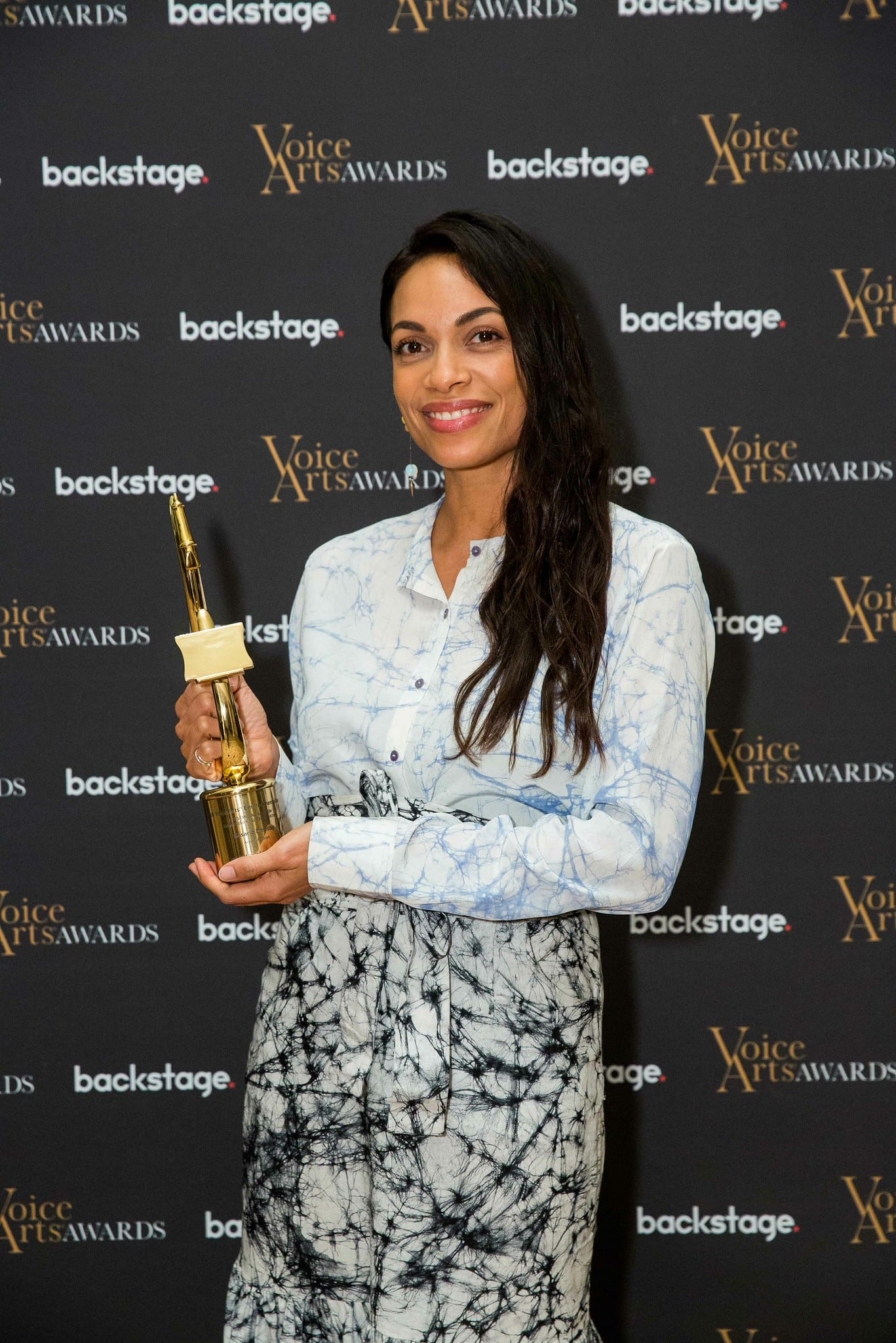Rosario Dawson holding a gold Voice Arts Award trophy in front of a Voice Arts Awards and Backstage step-and-repeat
