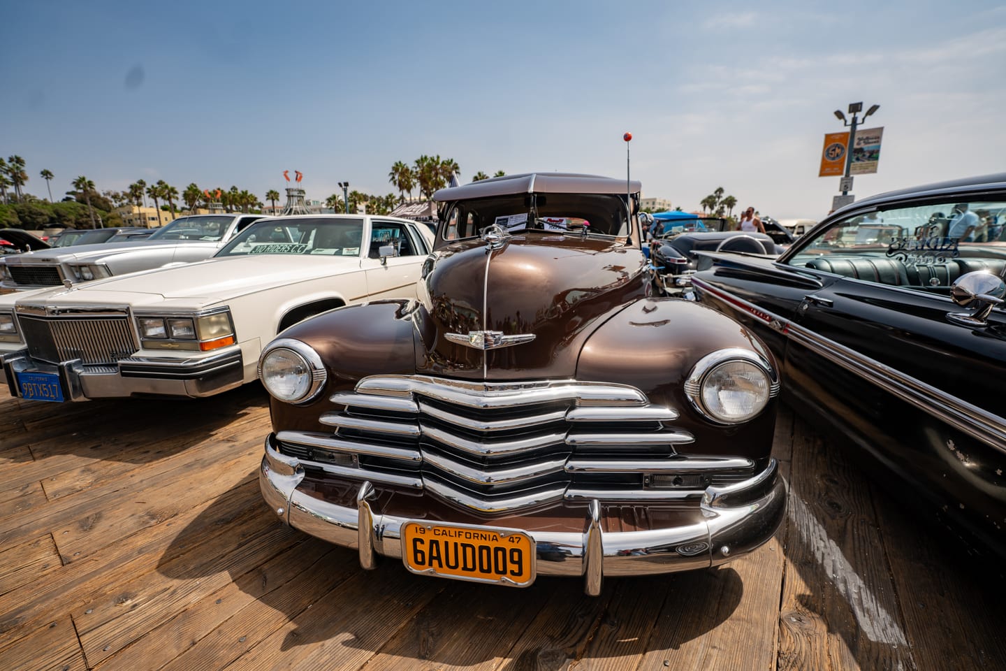 A brown 1947 Chevrolet with a heavy chrome grille and a yellow California plate parked on the wooden boards of the Santa Monica Pier at a classic car show, a white Cadillac and a black Impala either side and palm trees behind