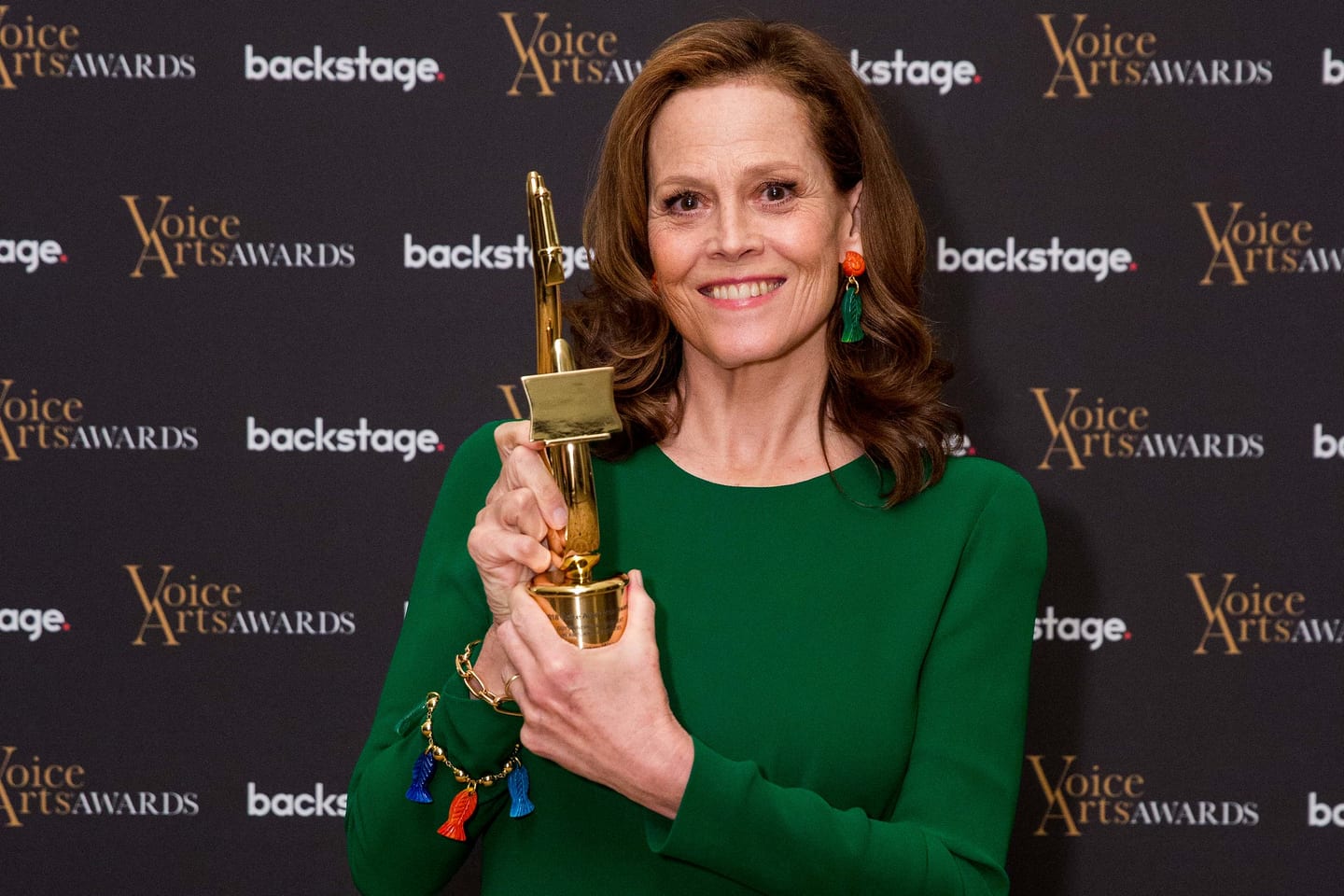 Sigourney Weaver in a green dress holding a gold Voice Arts Award trophy in front of a Voice Arts Awards and Backstage step-and-repeat