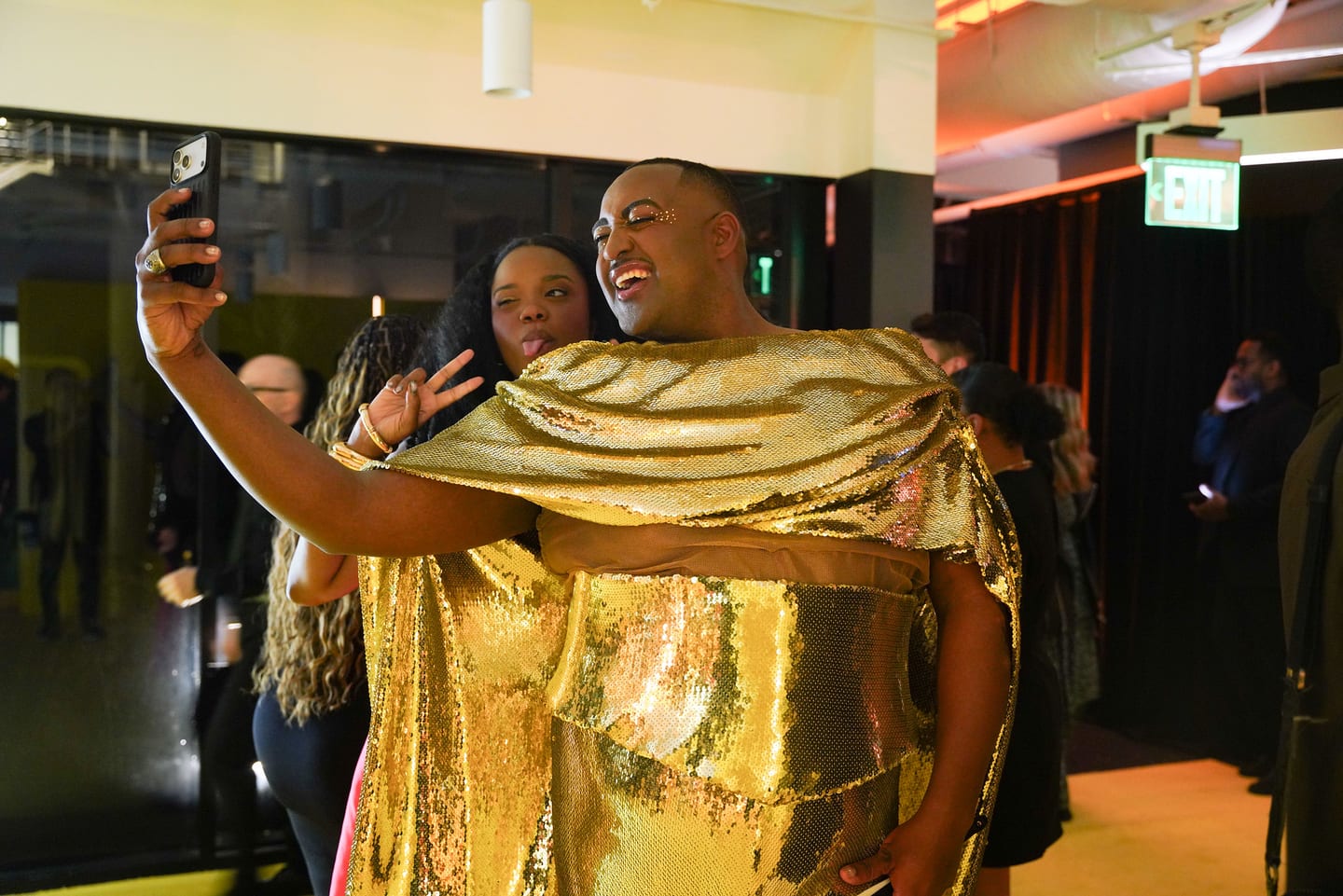 A guest in a gold cape taking a selfie with others at the after-party