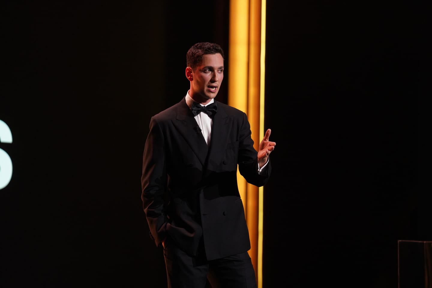 The host in a black tuxedo on stage between gold columns