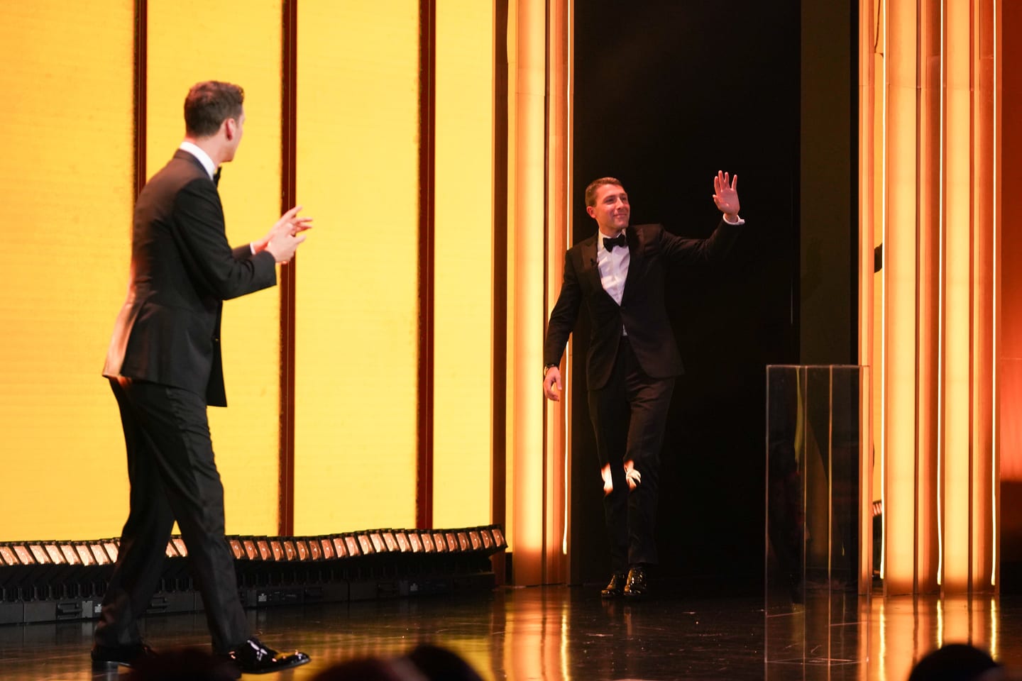 A presenter walking on stage toward the host against a gold striped backdrop