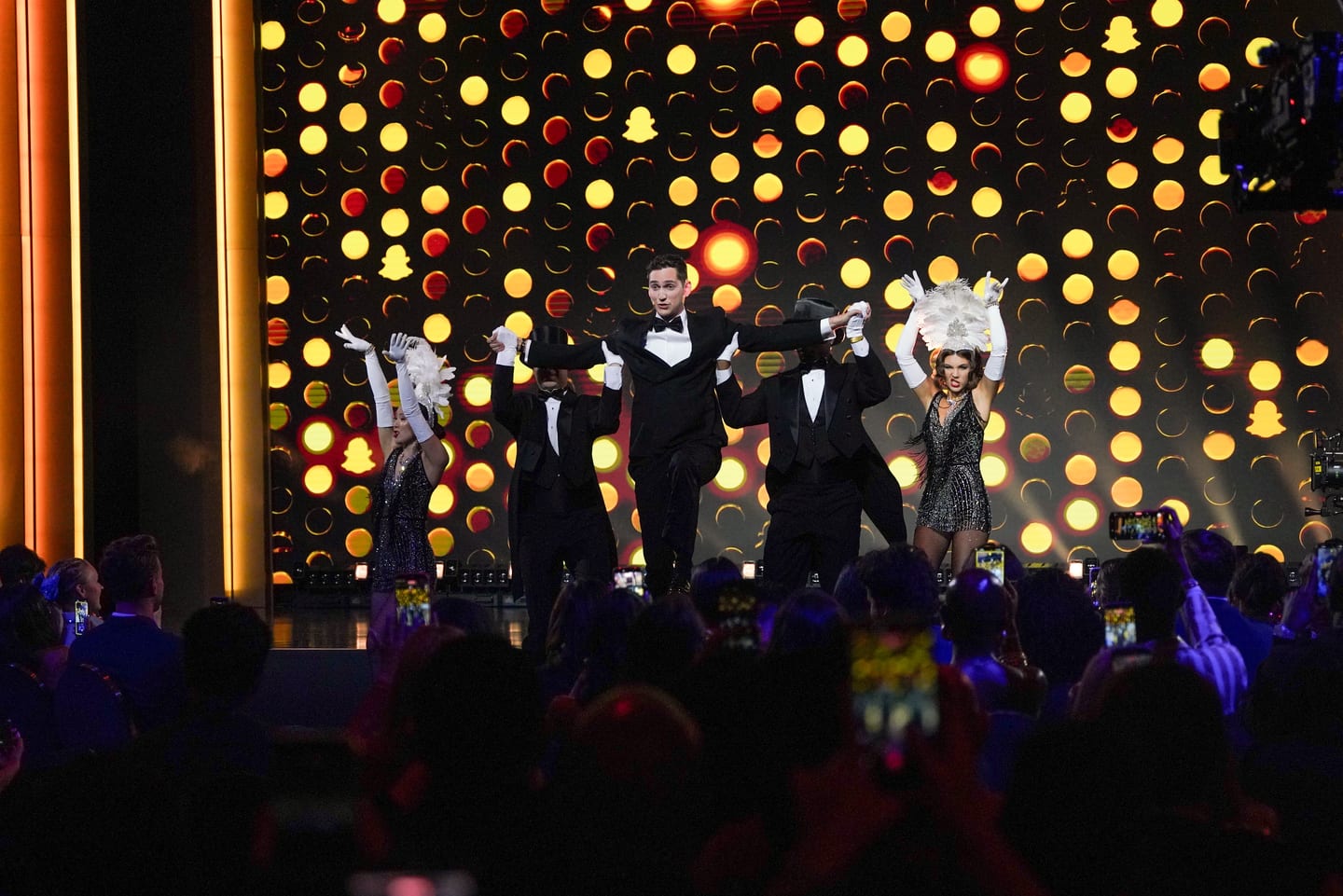 The full group on stage in front of a wall of coloured lights