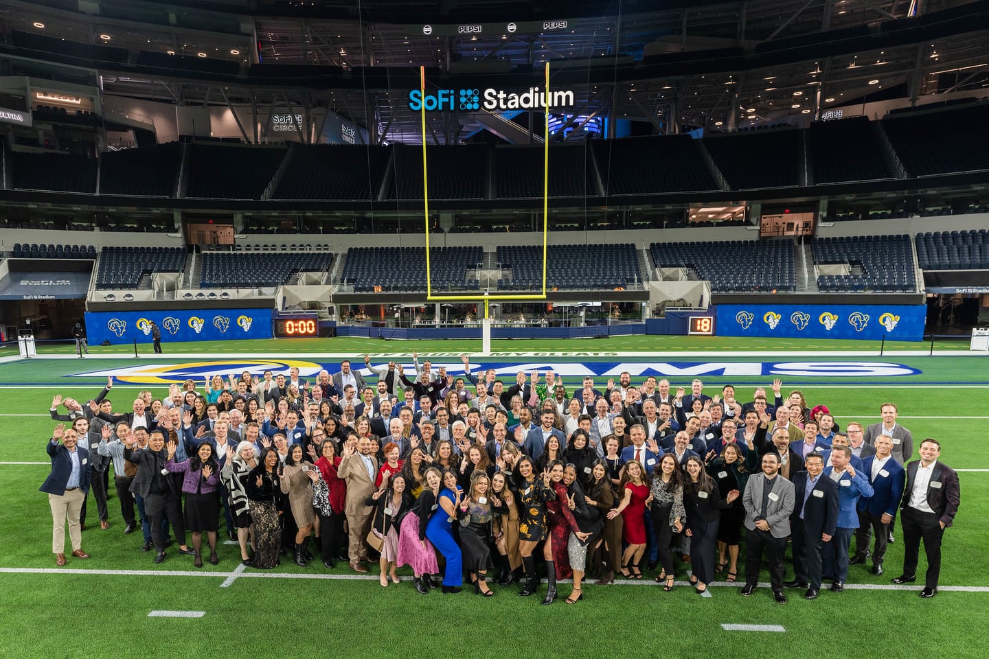 A large corporate group waving together on the field at SoFi Stadium with the stadium name and Rams branding behind them