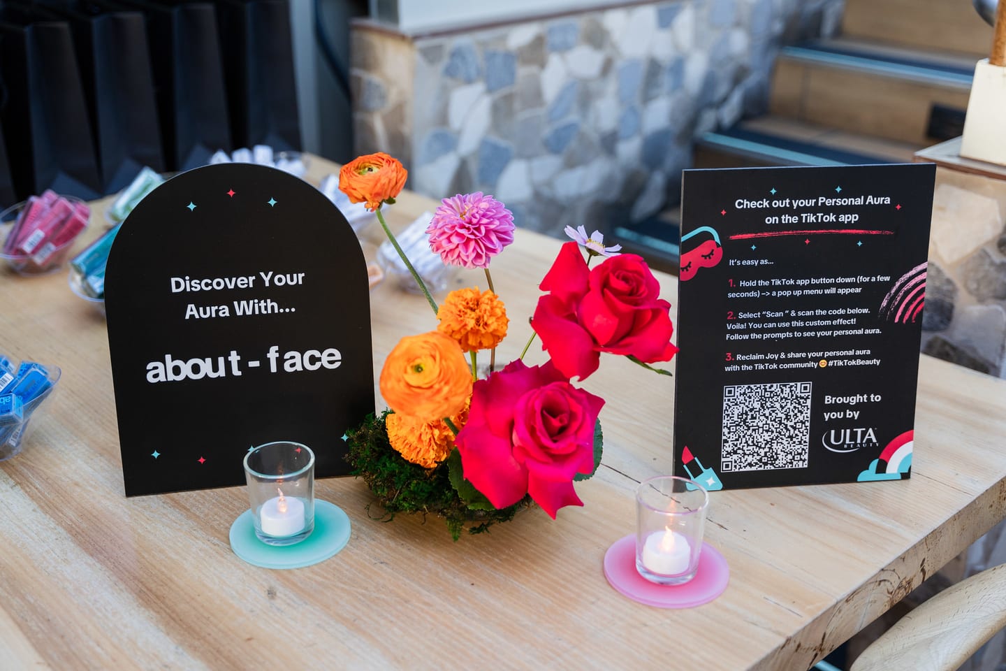 A black table sign reading Discover Your Aura With about-face, beside a QR card and bright florals at the TikTok and Ulta Beauty #TikTokBeauty dinner on the LouLou rooftop in Santa Monica
