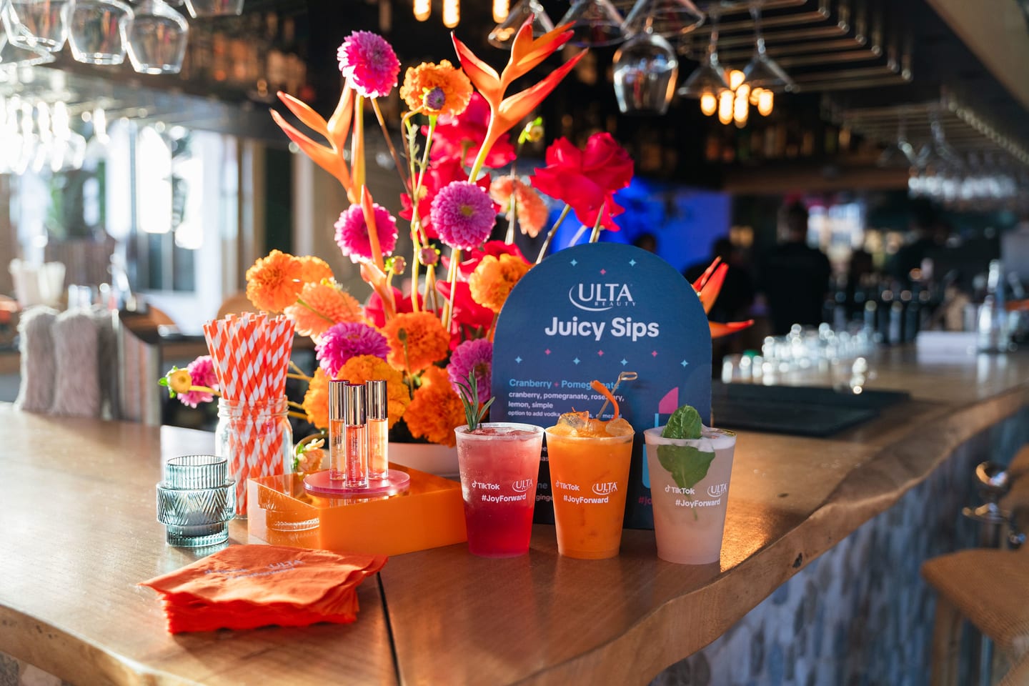 Cocktails lined up on a wooden bar beside an Ulta Beauty Juicy Sips menu card and a tall floral arrangement at the TikTok and Ulta Beauty #TikTokBeauty dinner on the LouLou rooftop in Santa Monica