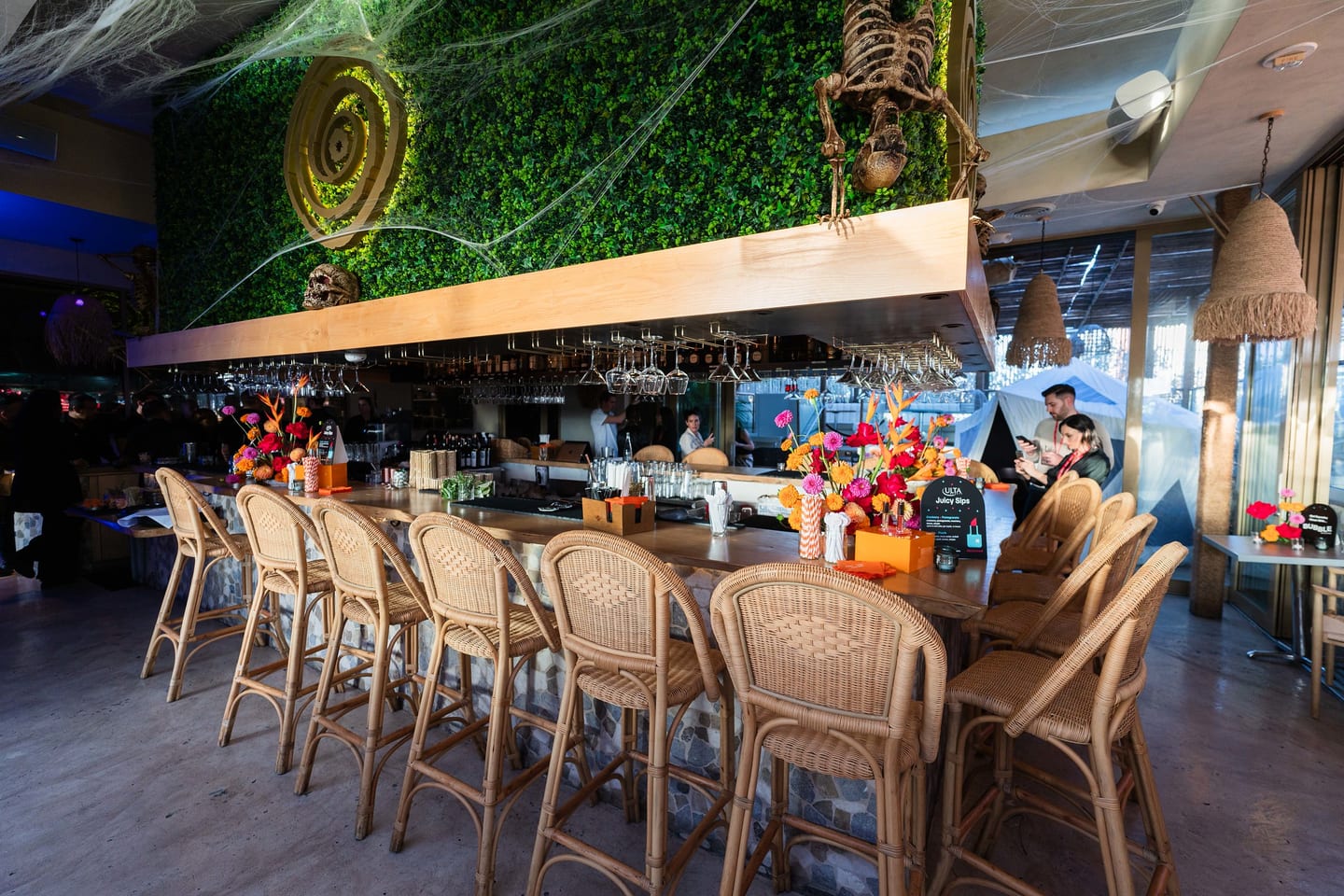 An empty rooftop bar lined with rattan stools and bright floral arrangements against a hedge wall at the TikTok and Ulta Beauty #TikTokBeauty dinner on the LouLou rooftop in Santa Monica