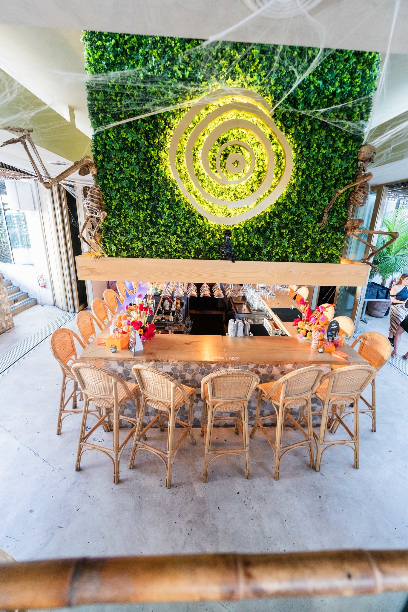 Rattan chairs and a bar set against a green hedge wall with a gold spiral logo on the LouLou rooftop, photographed before guests arrived at the TikTok and Ulta Beauty #TikTokBeauty dinner on the LouLou rooftop in Santa Monica