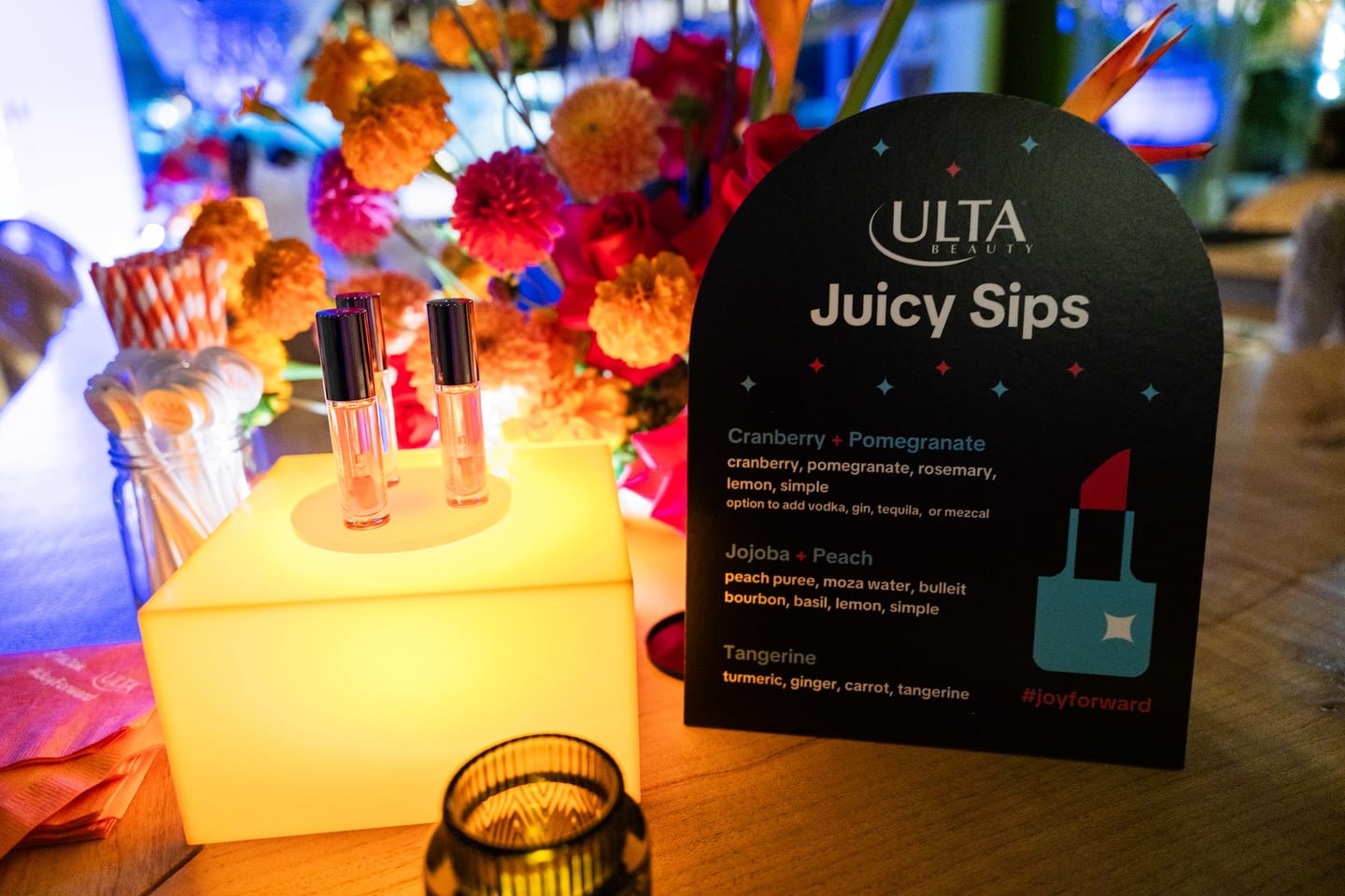 Lip products and an Ulta Beauty Juicy Sips menu card arranged on an illuminated orange cube among marigolds at the TikTok and Ulta Beauty #TikTokBeauty dinner on the LouLou rooftop in Santa Monica