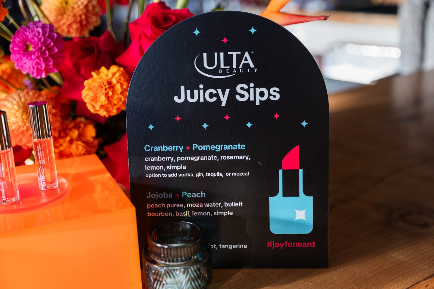 An Ulta Beauty Juicy Sips cocktail menu card printed with a lipstick illustration, standing among bright florals at the TikTok and Ulta Beauty #TikTokBeauty dinner on the LouLou rooftop in Santa Monica