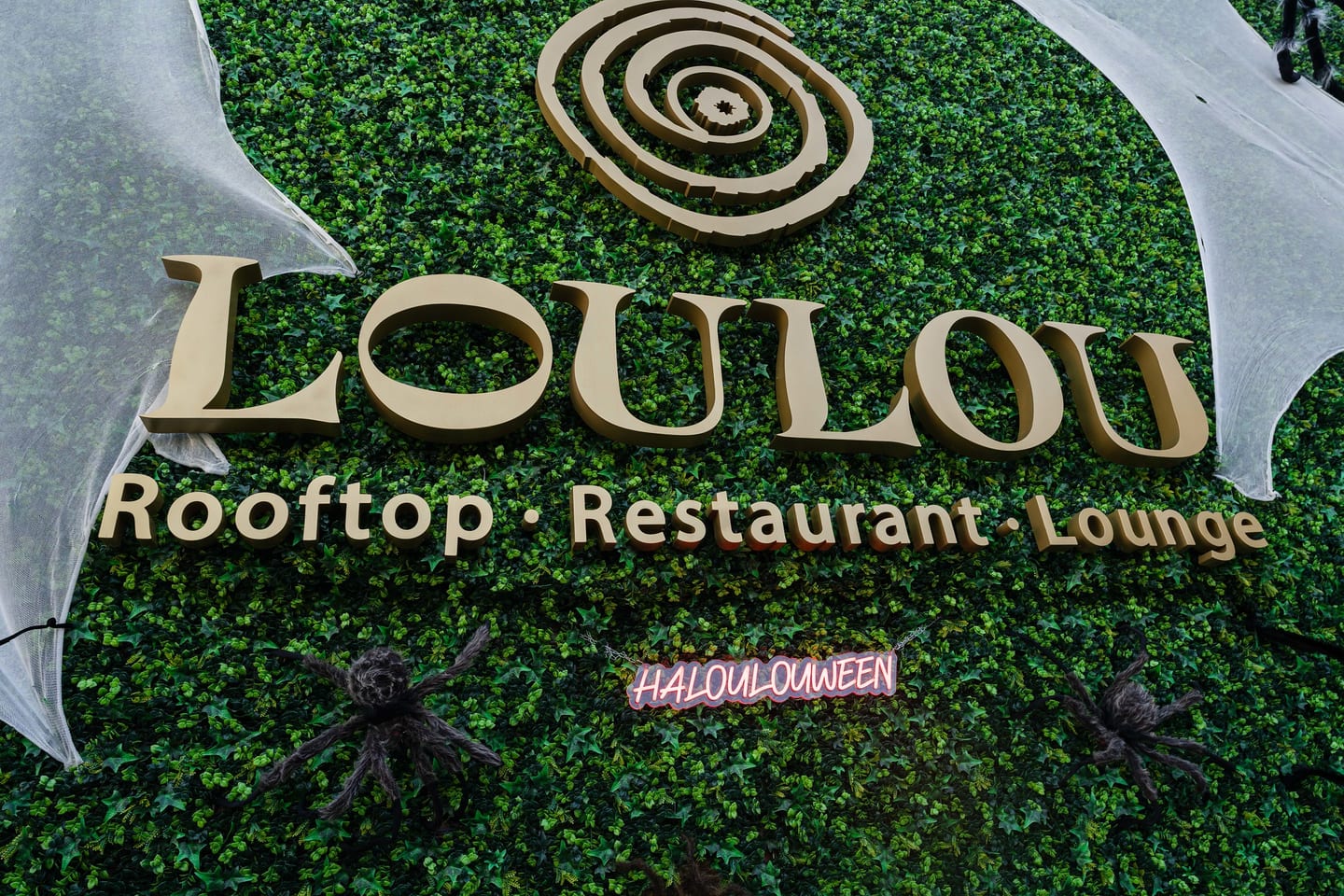 A gold LouLou Rooftop Restaurant Lounge sign mounted on a green hedge wall, dressed with Halloween cobwebs and spiders and a pink HALOULOUWEEN neon, at the entrance to a TikTok and Ulta Beauty event in Santa Monica