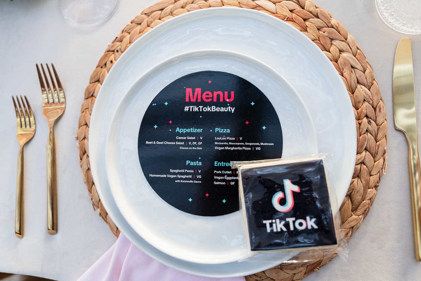 An overhead place setting with a round #TikTokBeauty menu disc on a white plate over a woven charger, gold flatware and a TikTok coaster at the TikTok and Ulta Beauty #TikTokBeauty dinner on the LouLou rooftop in Santa Monica