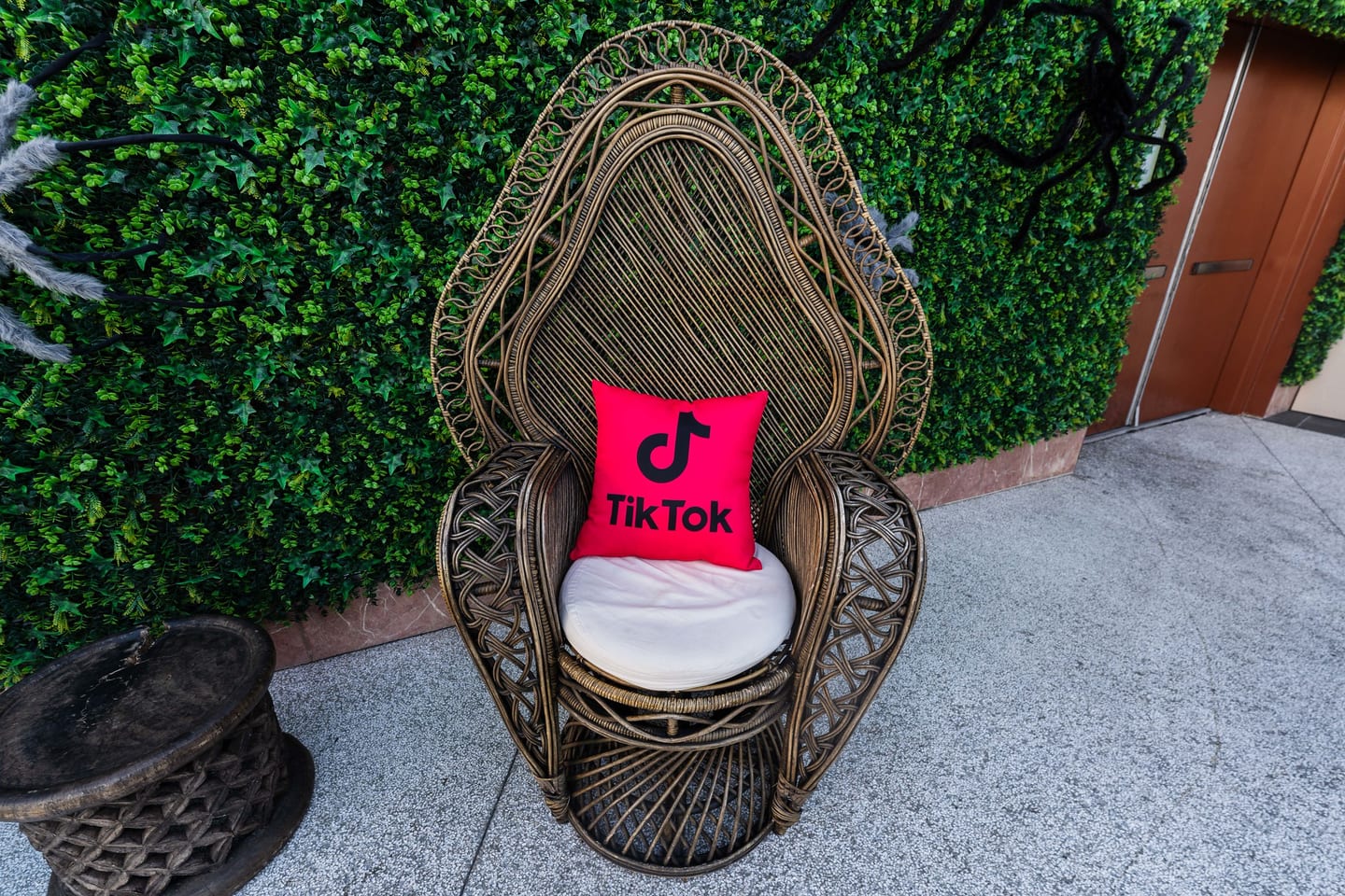 A rattan peacock chair holding a pink TikTok logo pillow, staged against a green hedge wall at the TikTok and Ulta Beauty #TikTokBeauty dinner on the LouLou rooftop in Santa Monica