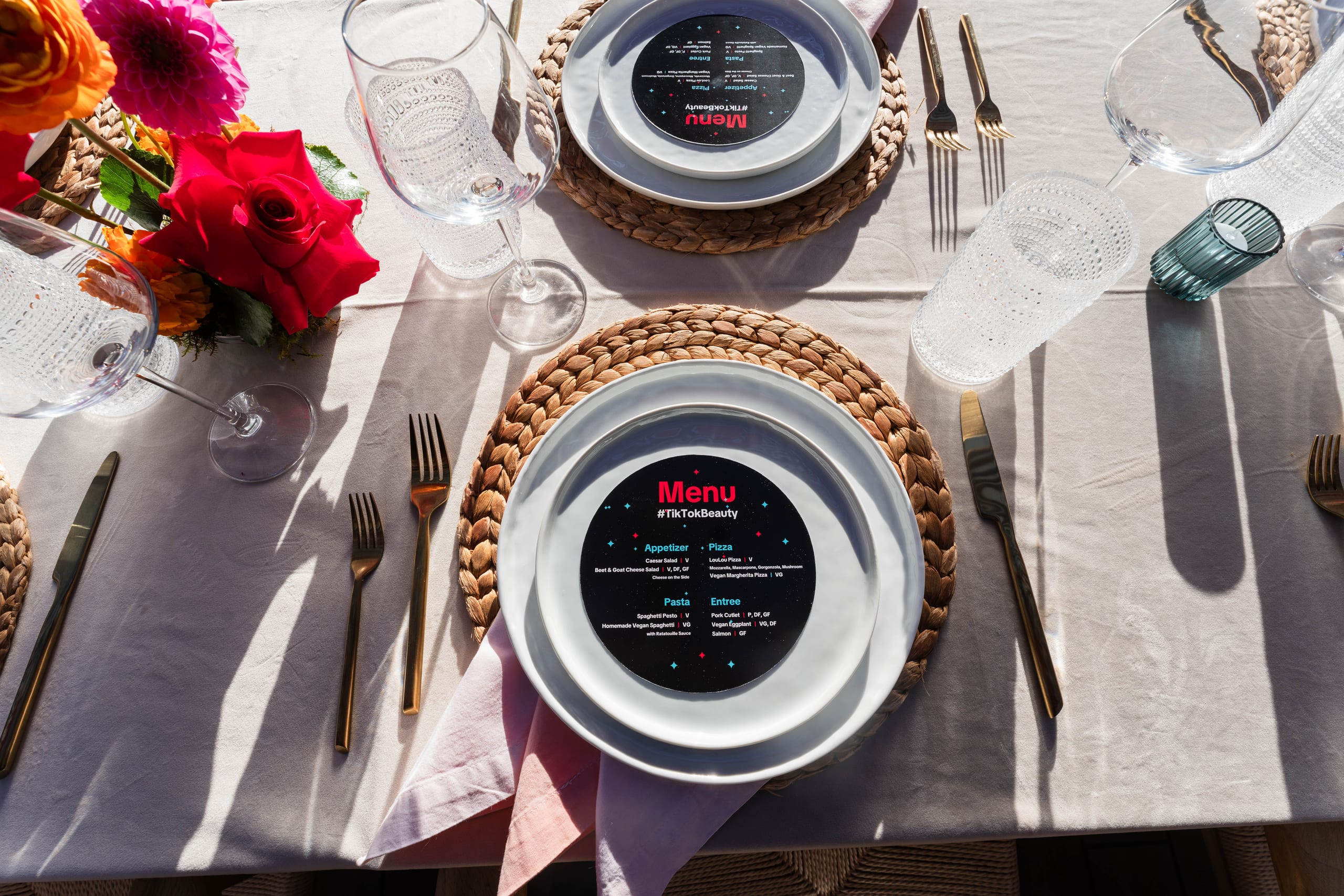 An overhead place setting with a branded #TikTokBeauty menu card on a white plate over a rattan charger, gold flatware, textured glassware and roses at a TikTok Beauty brand dinner in Los Angeles