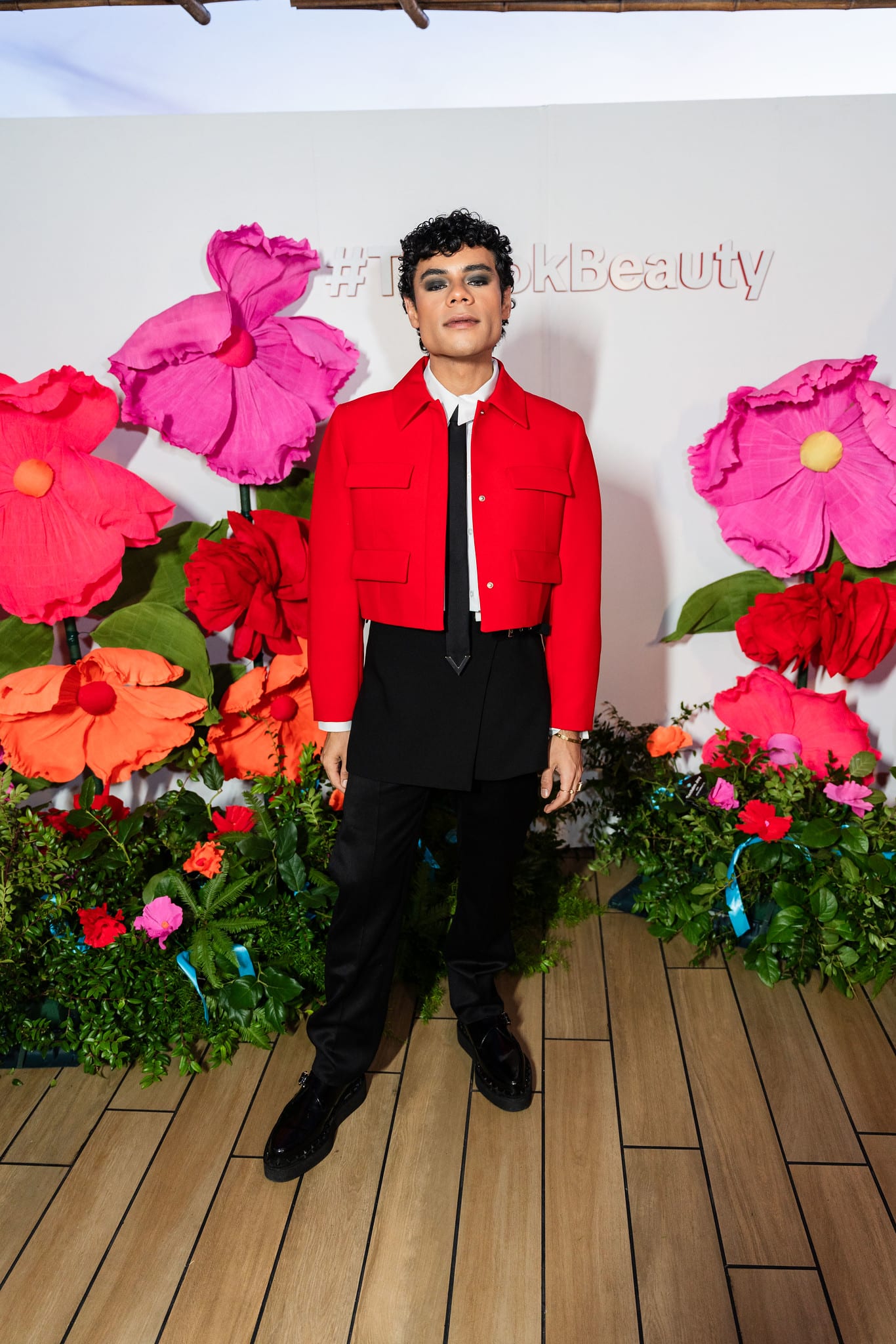 A guest in a red jacket posing at the #TikTokBeauty step-and-repeat built from oversized paper flowers at the TikTok and Ulta Beauty #TikTokBeauty dinner on the LouLou rooftop in Santa Monica