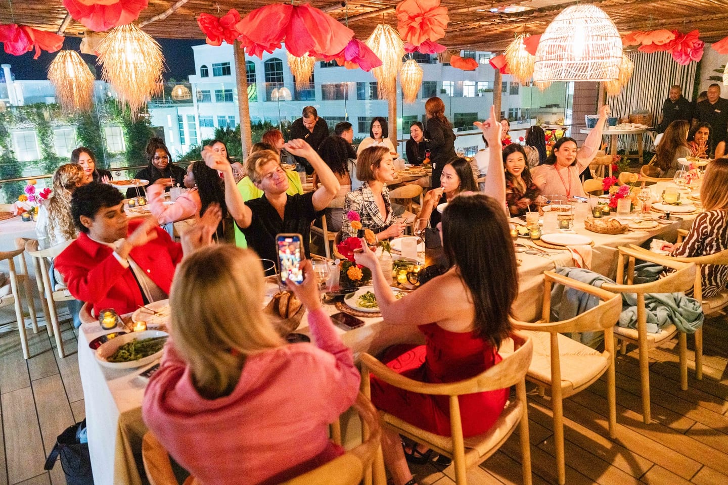 Guests seated along the full length of the communal rooftop table at LouLou in Santa Monica, hands raised mid-toast beneath oversized paper flowers and woven pendant lights, at the TikTok and Ulta Beauty #TikTokBeauty dinner