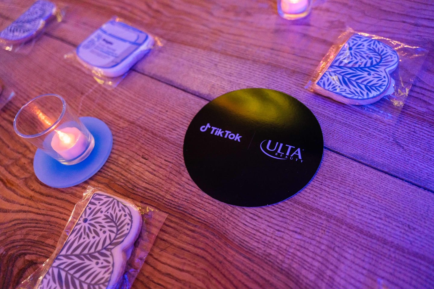 A black coaster printed with the TikTok and Ulta Beauty logos on a wooden table lit purple, beside candles at the TikTok and Ulta Beauty #TikTokBeauty dinner on the LouLou rooftop in Santa Monica