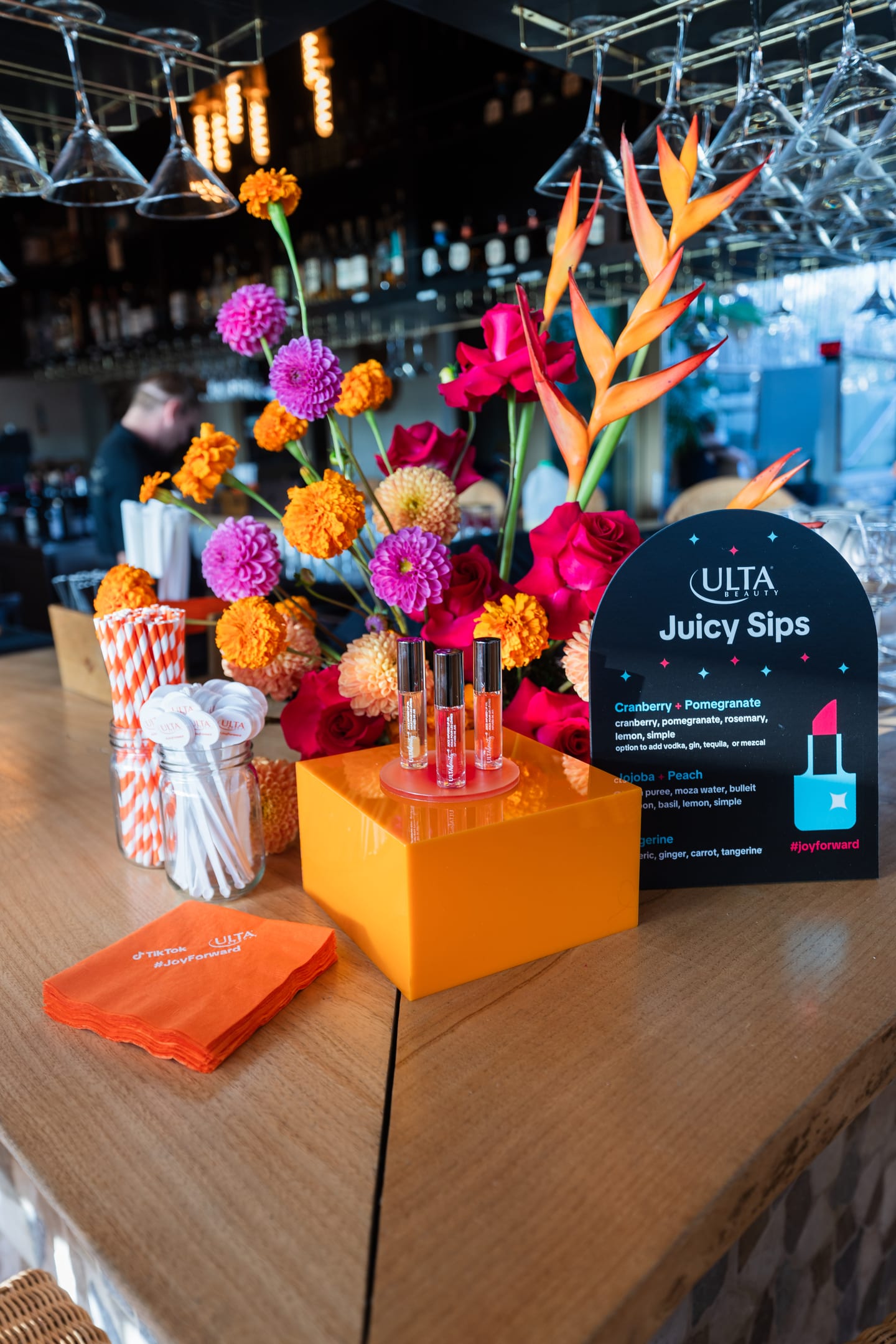An Ulta Beauty “Juicy Sips” lip-oil product display with bright florals and TikTok #JoyForward napkins at a TikTok beauty activation in Los Angeles