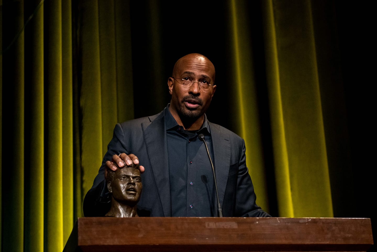 Van Jones speaking at a wooden podium with one hand resting on a bronze bust, a gold-lit stage curtain behind him