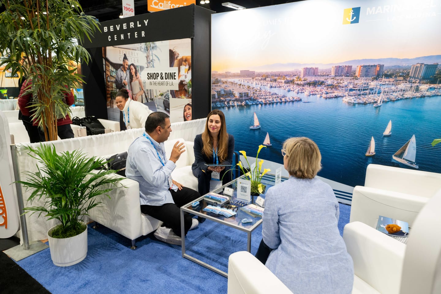 Attendees meeting on white lounge chairs in front of a marina backdrop at a California pavilion booth at IPW