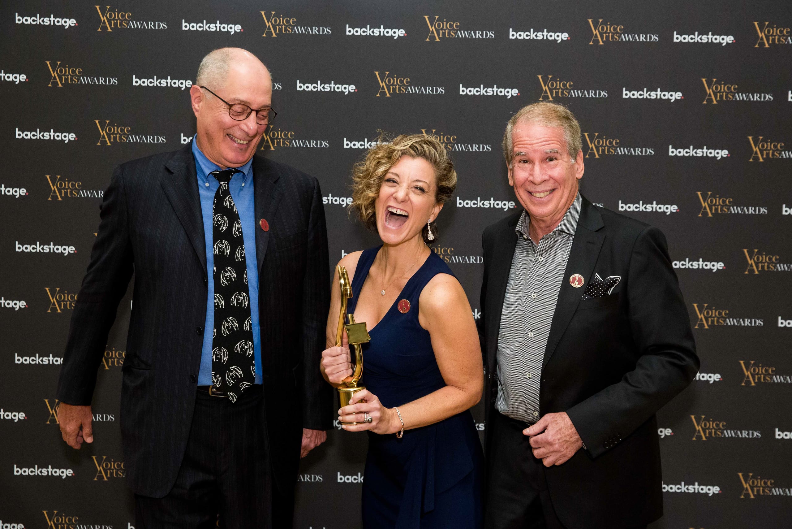 An award winner laughing while holding a gold trophy between two presenters on the Voice Arts Awards and Backstage step-and-repeat
